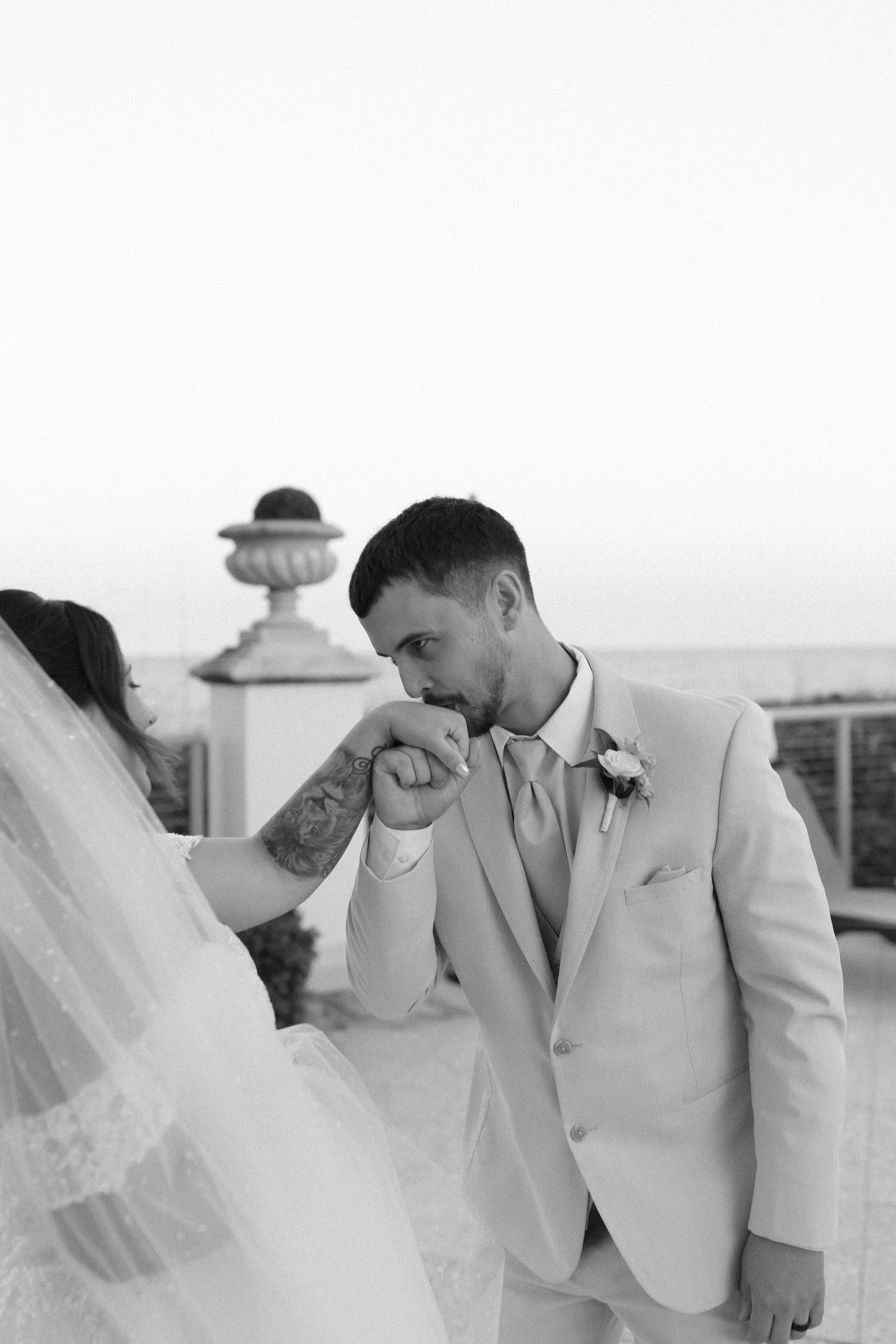 Groom kisses bride's hand in a tender black and white wedding portrait on an outdoor terrace.