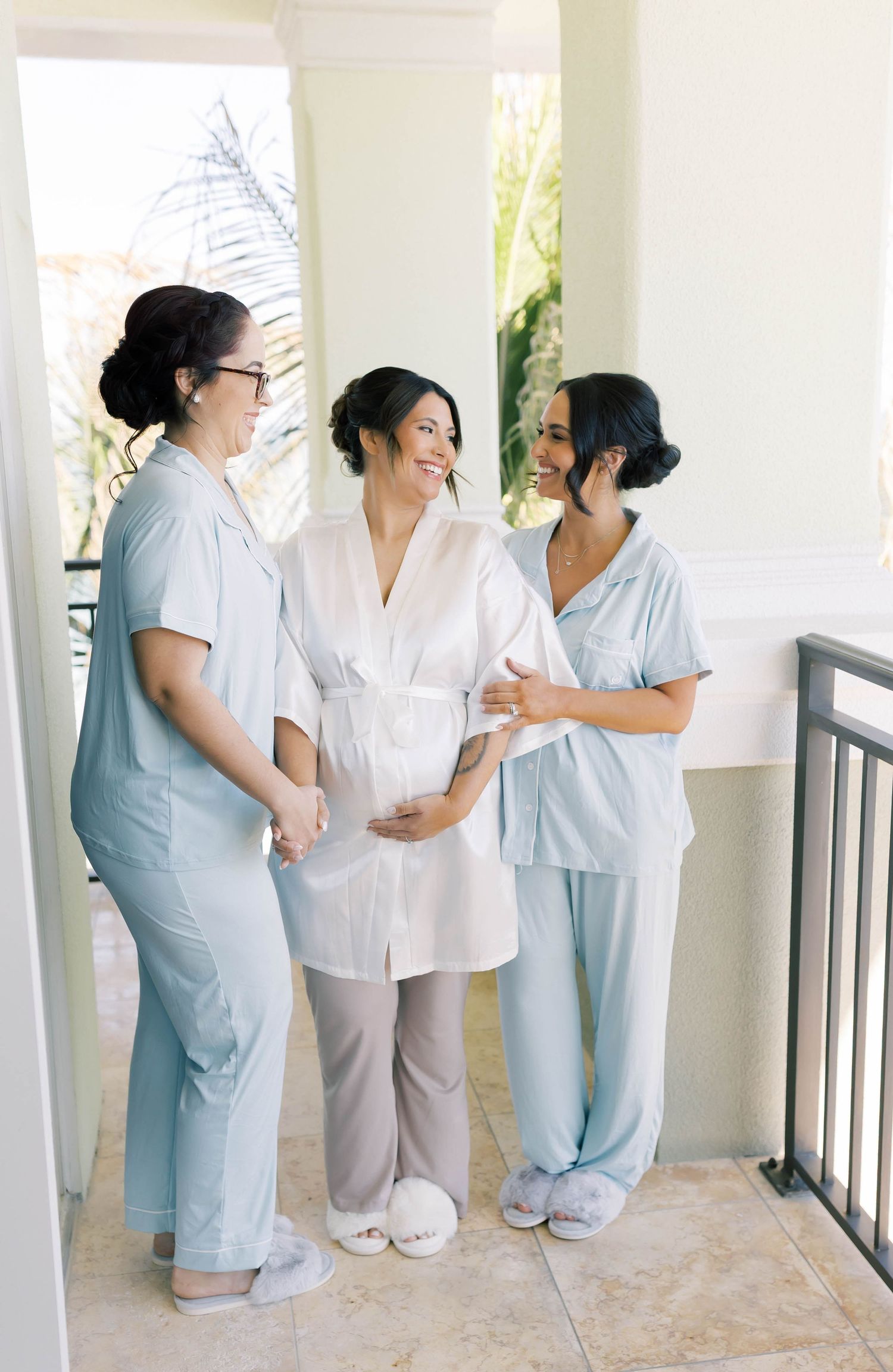 Pregnant bride in white robe with bridesmaids in light blue pajamas sharing a joyful moment on a balcony.