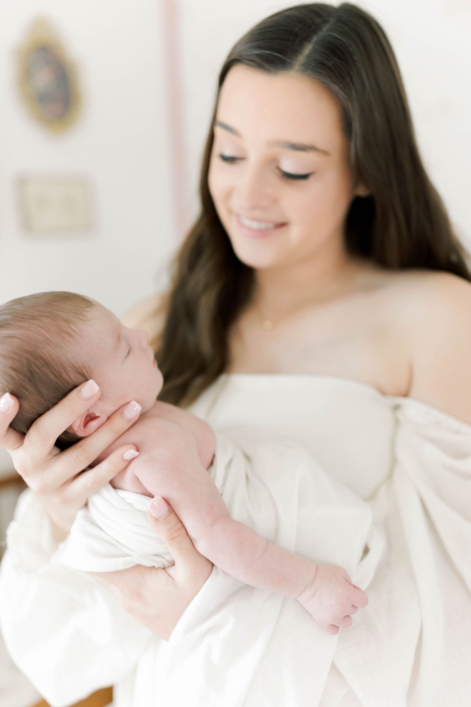 Tender moment between mother and newborn baby in white dress against light background.