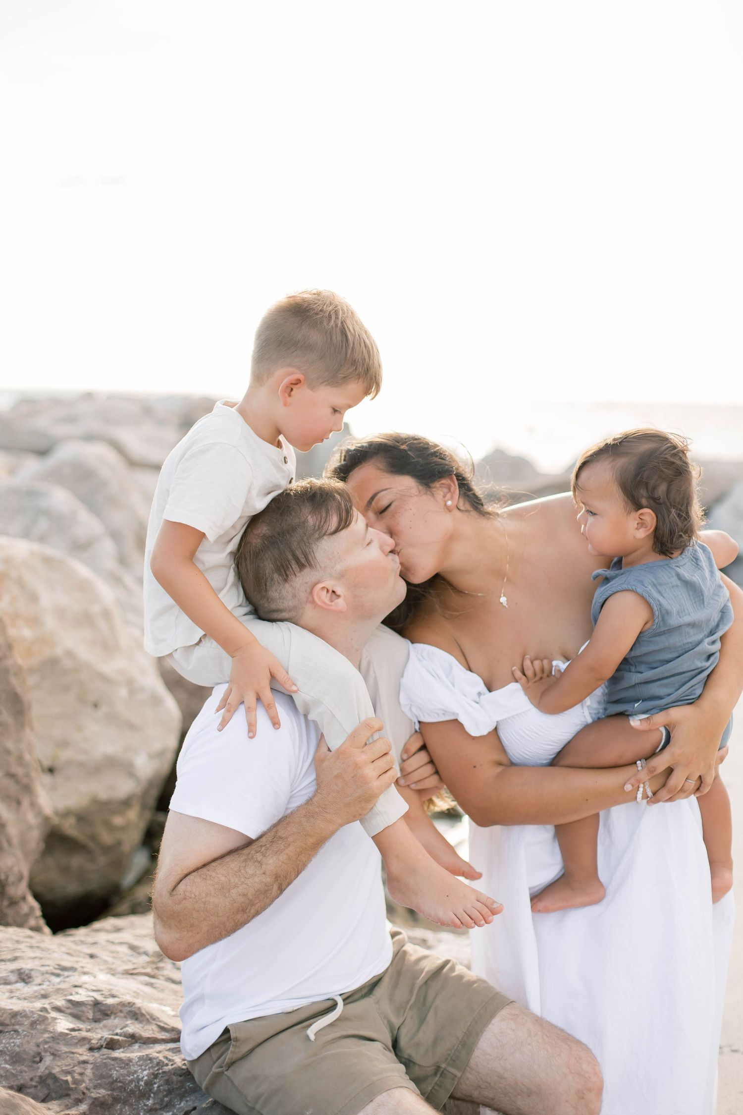 A loving family shares embraces and kisses on a sunny beach with rocky backdrop.