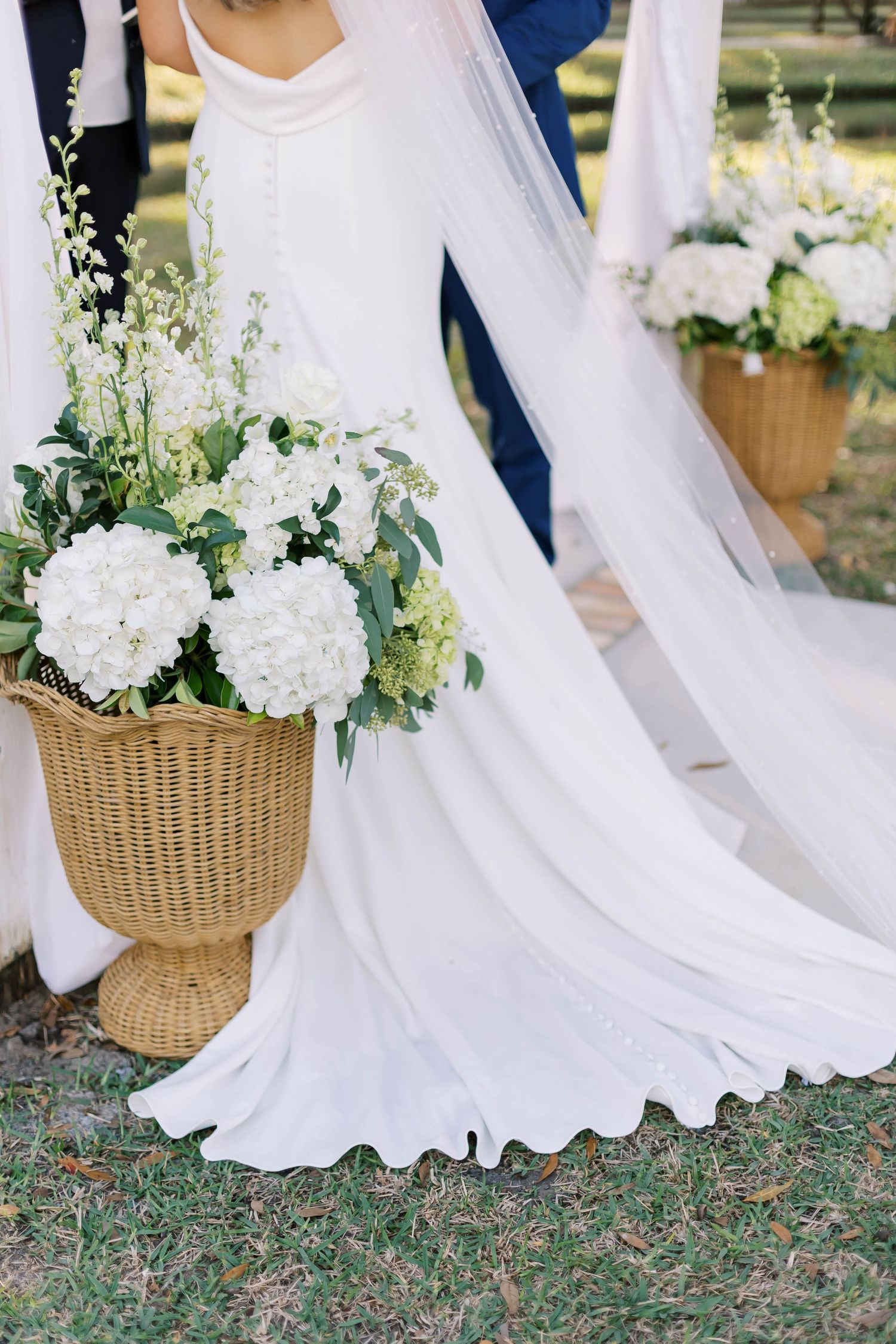 White fabric draping and woven baskets with flowers line an outdoor wedding aisle.