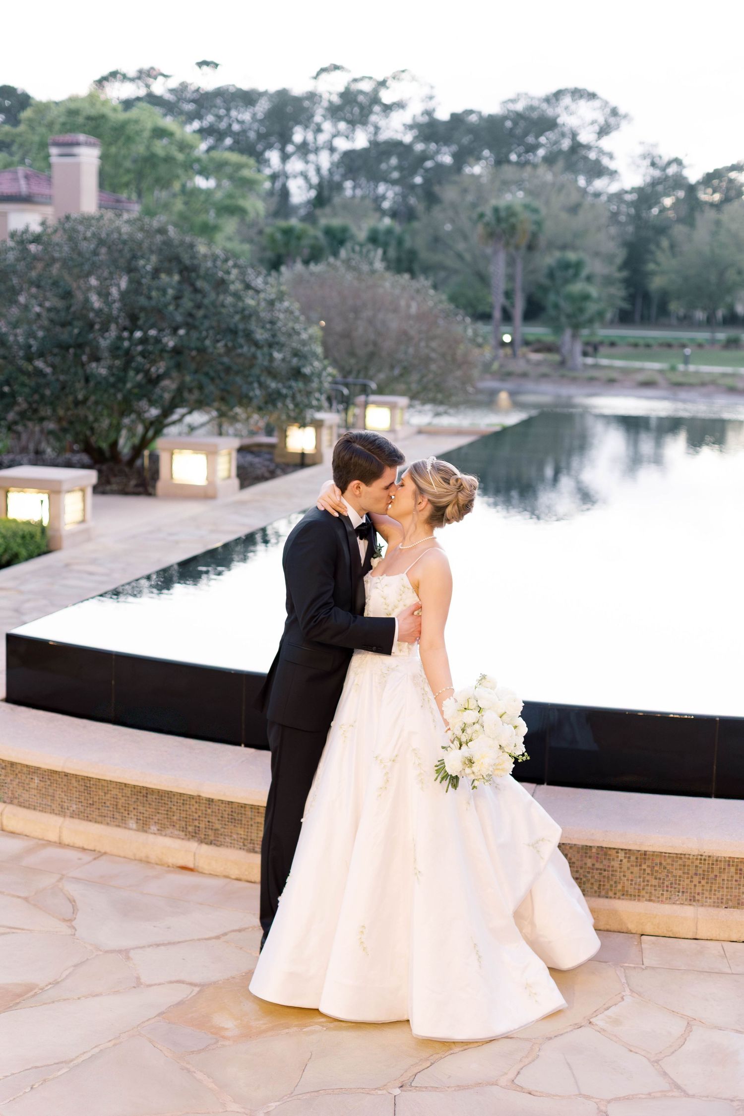 Bride and groom share a romantic kiss in elegant wedding attire beside a serene reflective pond at dusk.