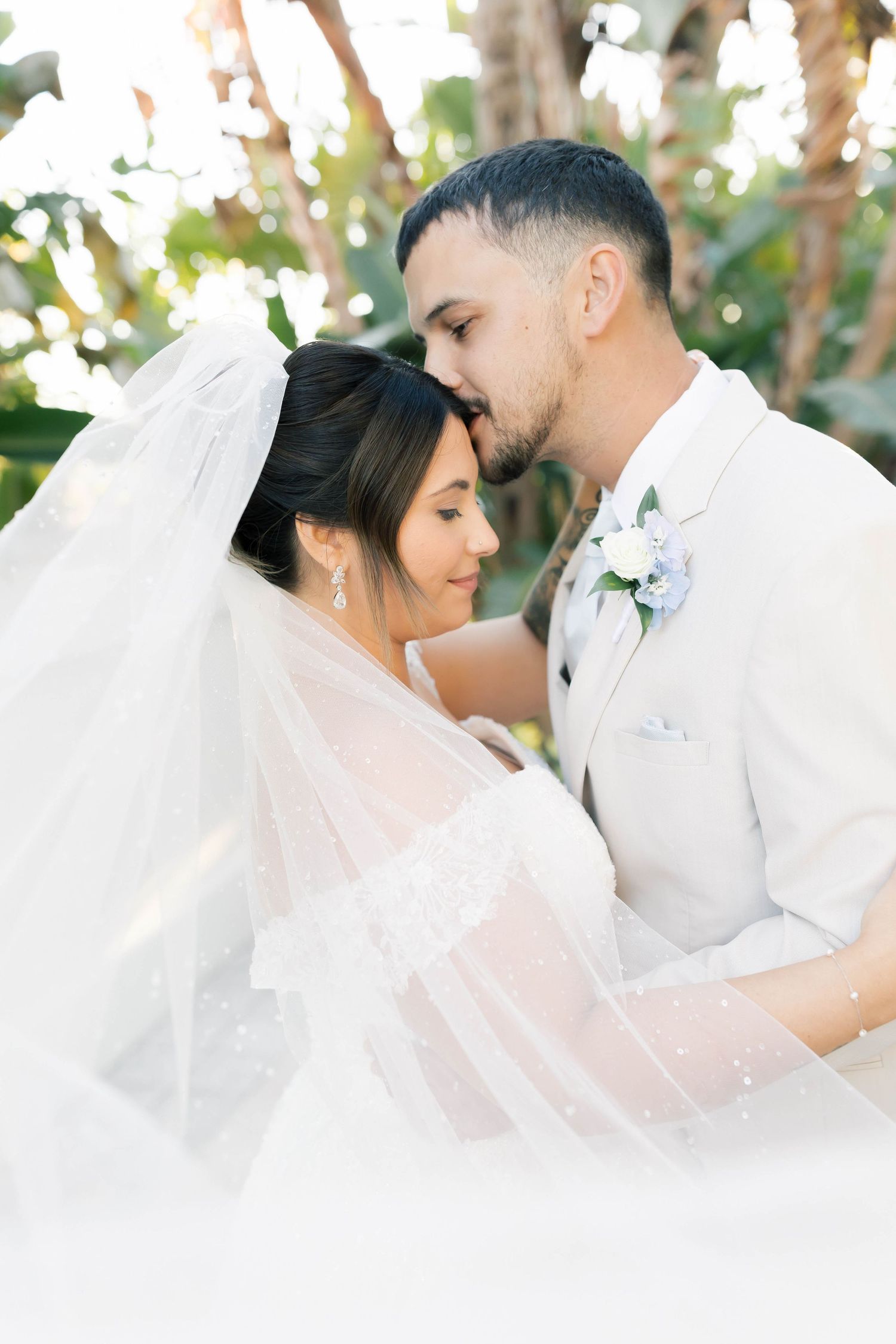 Bride and groom share an intimate moment, groom kissing bride's forehead, wrapped in a flowing veil outdoors.