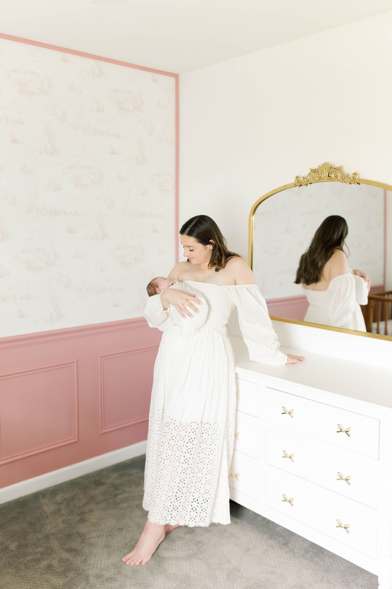 Someone in a white dress holds a newborn baby beside a gold-framed mirror in a nursery.