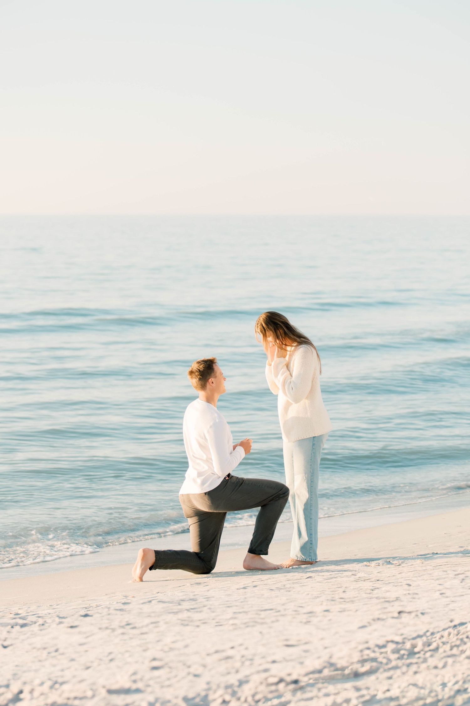 A couple kneels together on a pristine white sandy beach at sunset with calm ocean waters in the background.