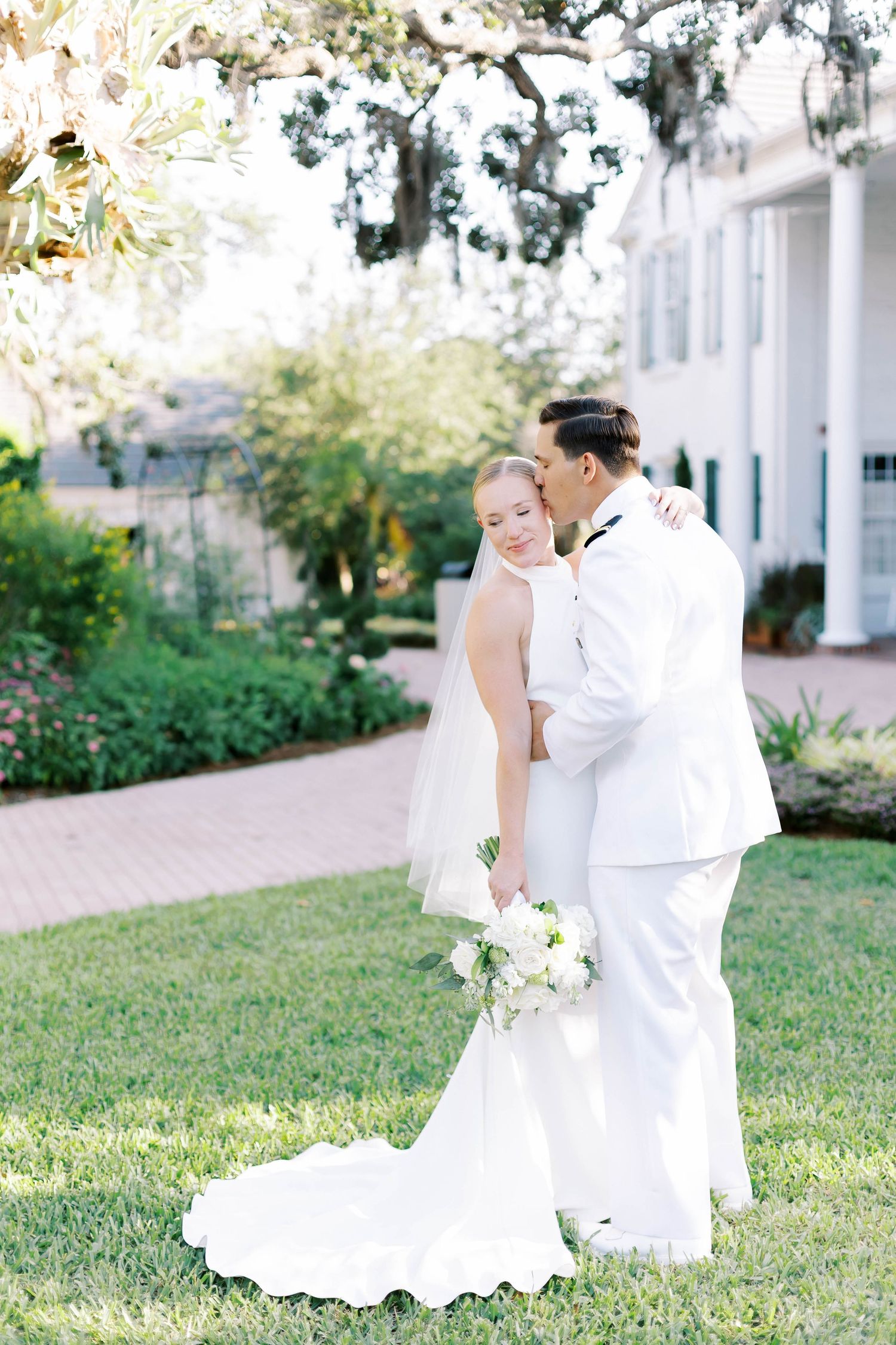 Bride and groom share a tender kiss outdoors on lush green lawn beside elegant white estate building.