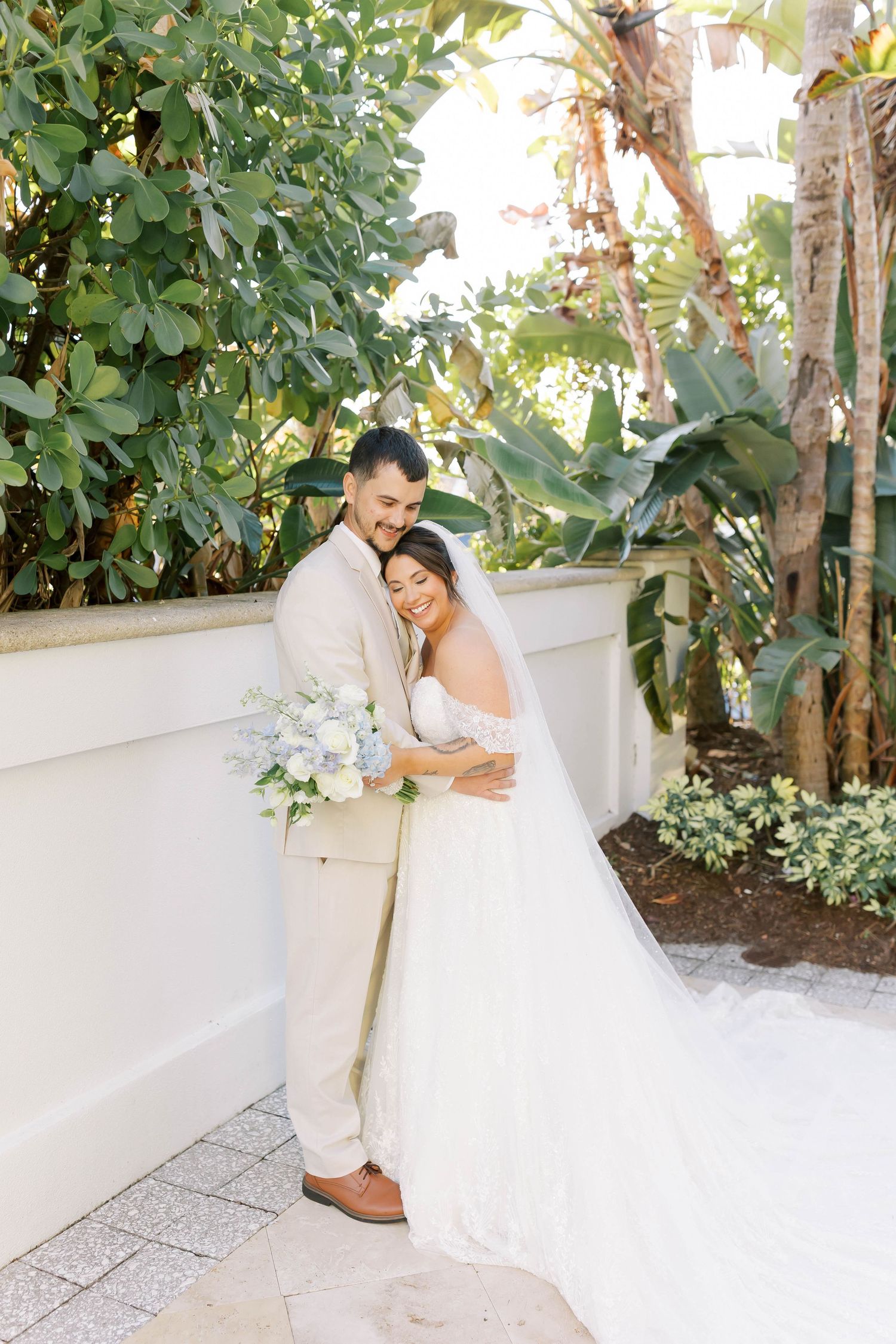 Groom embraces bride from behind as she smiles, both surrounded by lush tropical greenery.