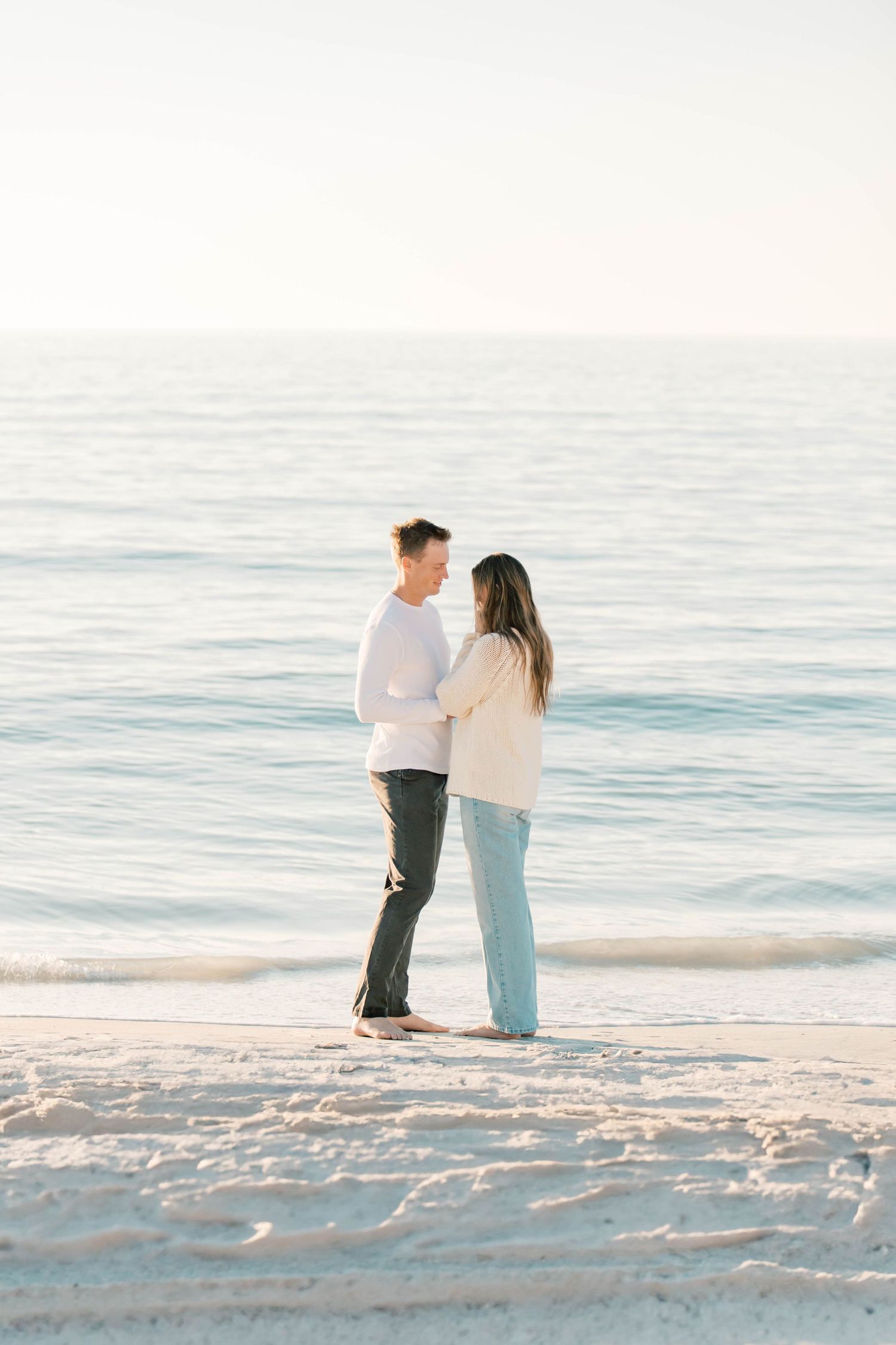 A couple shares an intimate moment while standing on a white sandy beach at sunset.