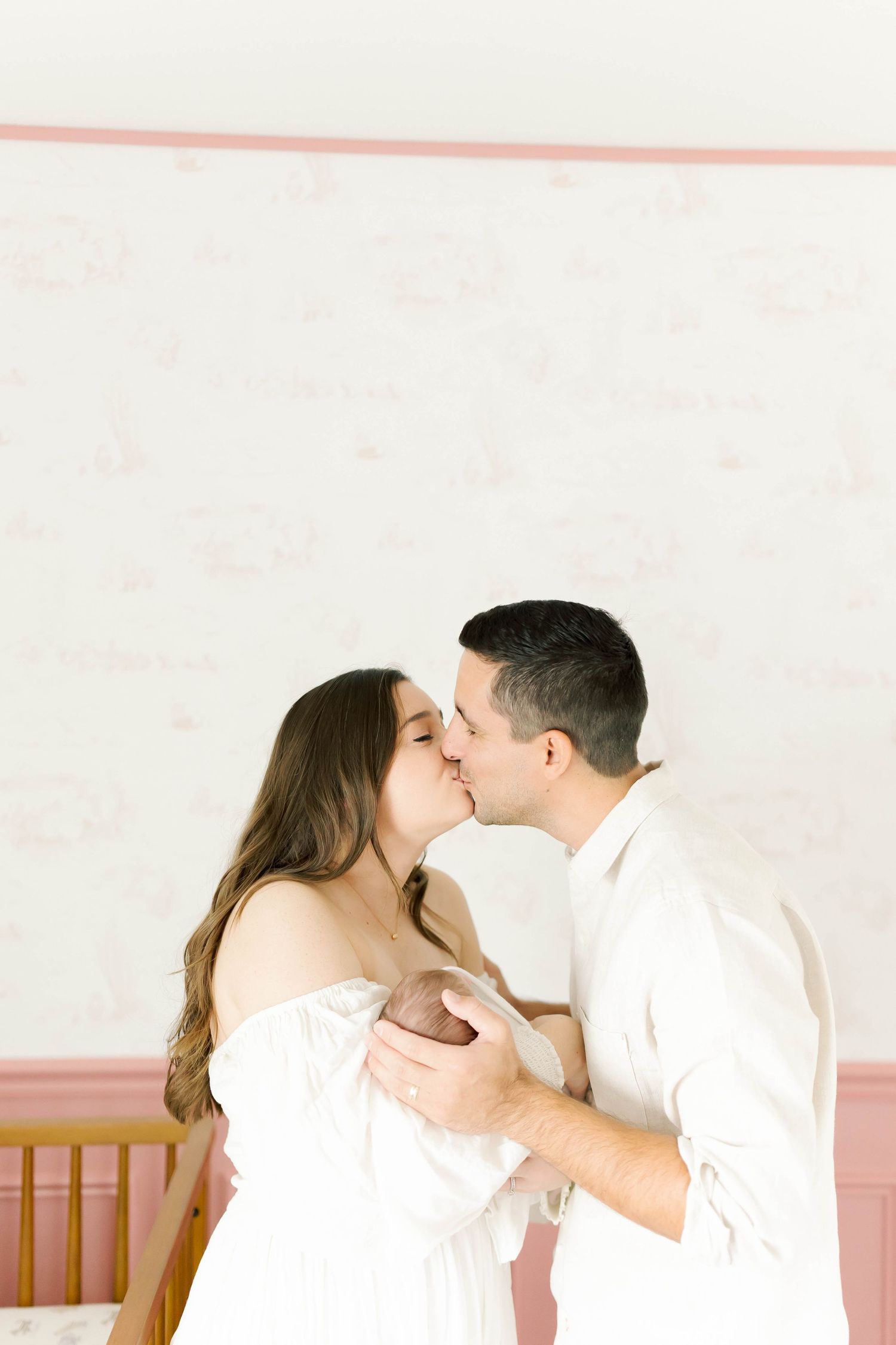 Romantic couple in white sharing a gentle kiss against a soft pink and white wall.