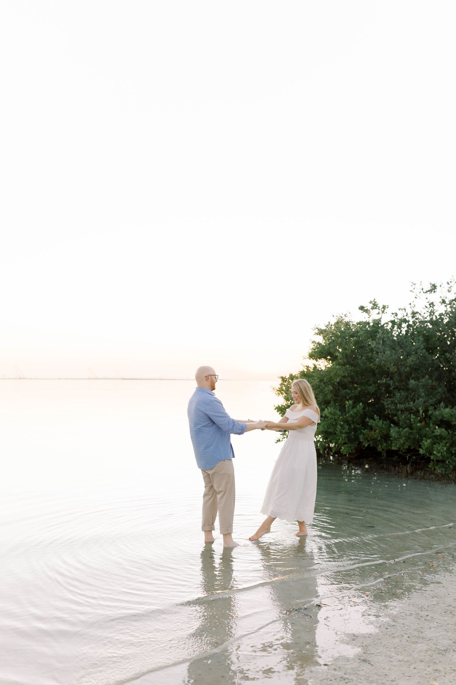 A couple holds hands while standing in shallow water near mangroves during sunset.