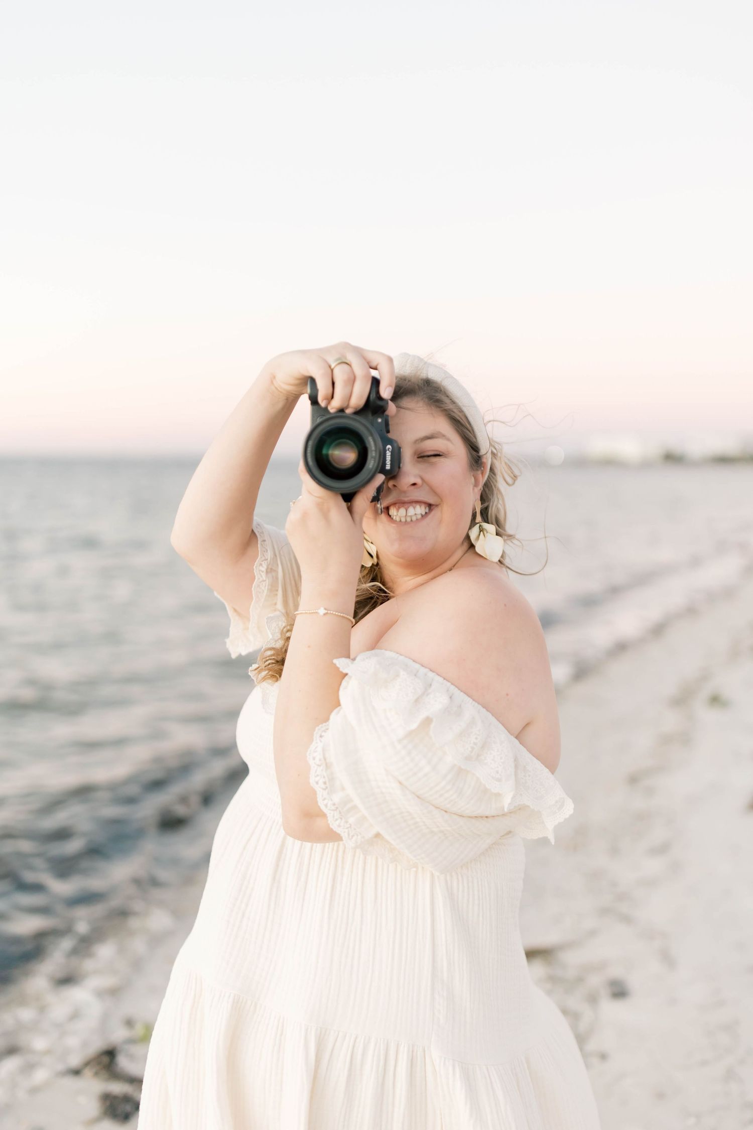 Woman in white off-shoulder dress smiling while holding DSLR camera on a beach at sunset.