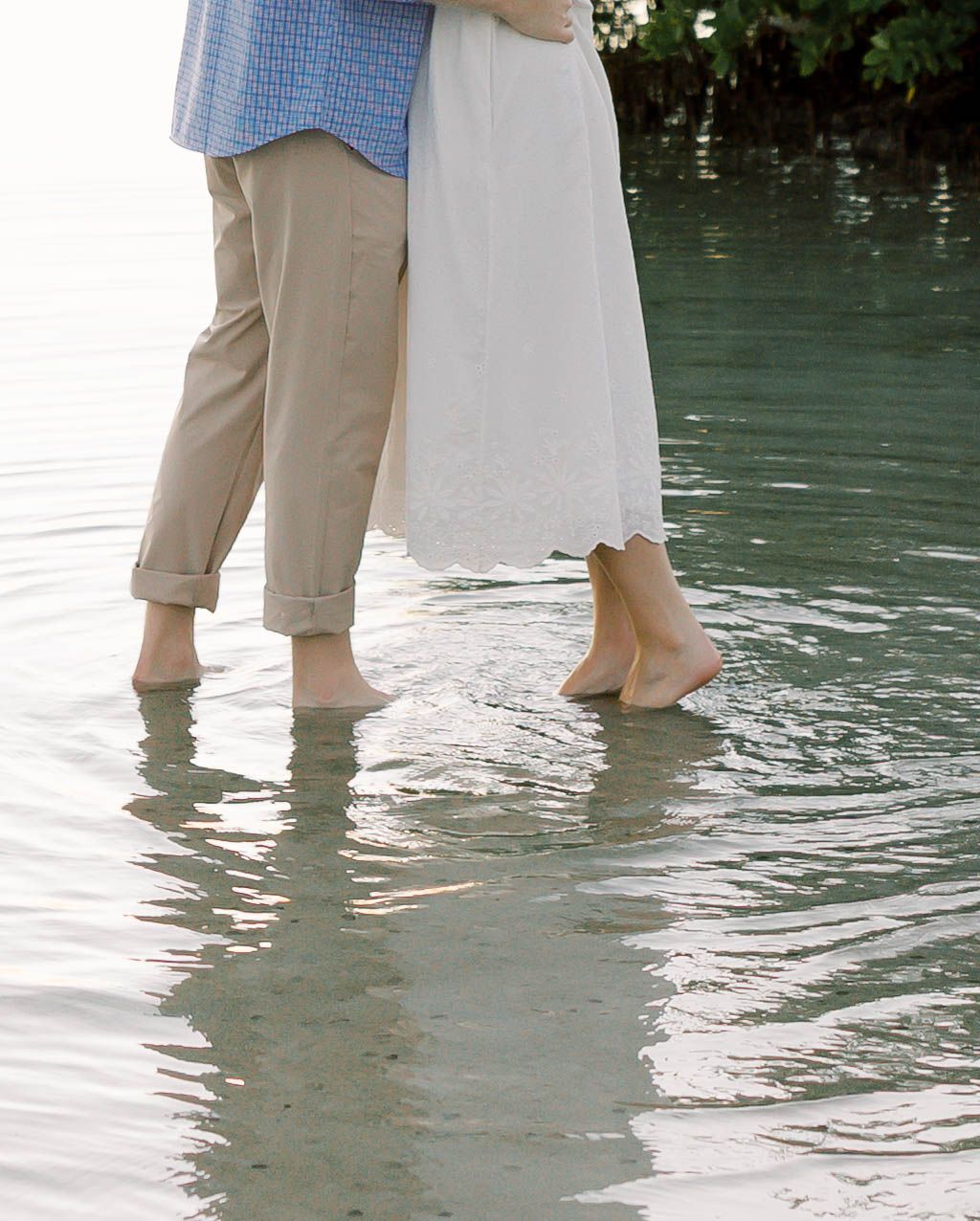 Cropped view of couple's feet standing in shallow ocean water wearing casual attire.