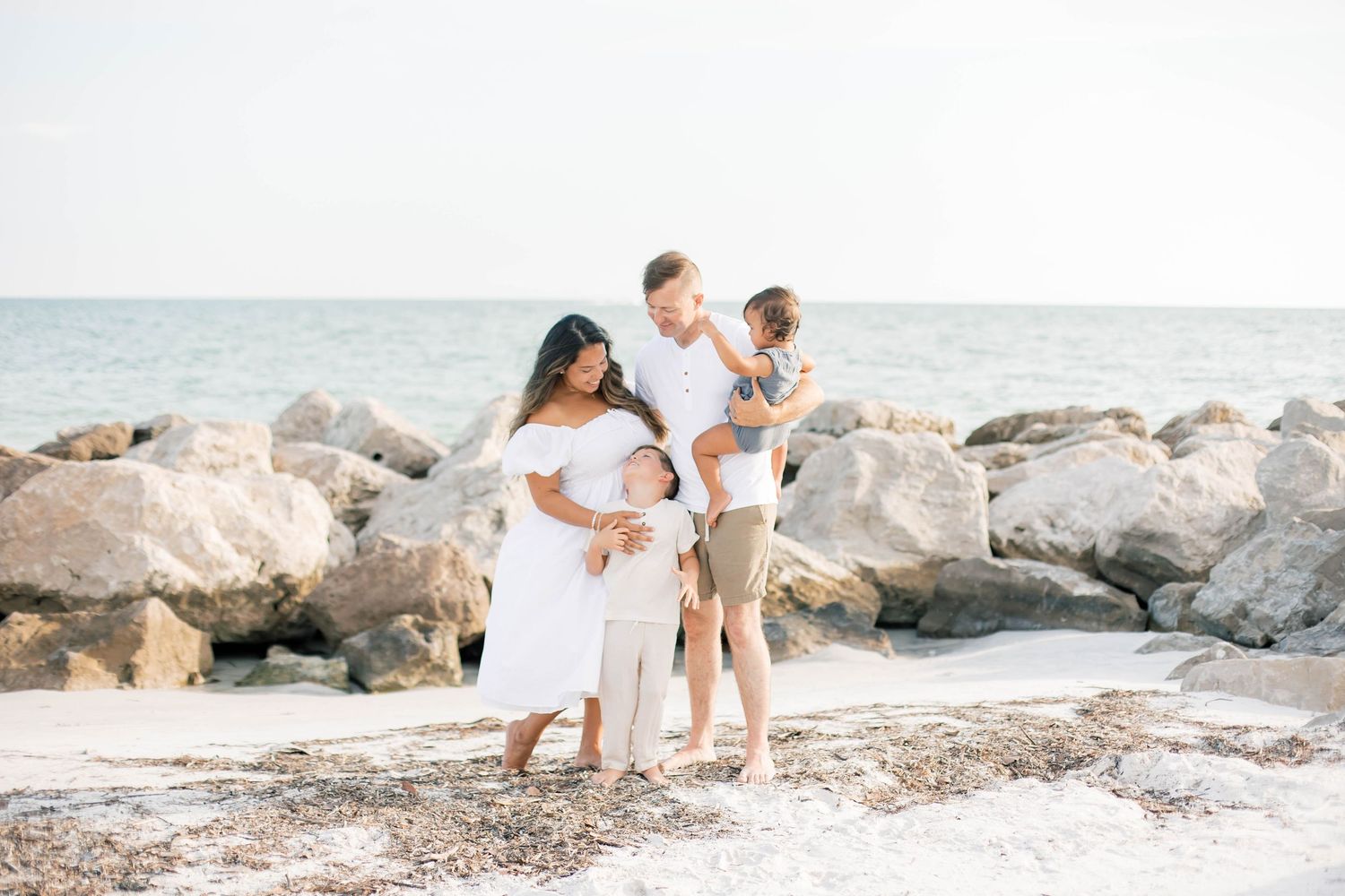A sweet family moment captured on a beach with large rocks and ocean backdrop.