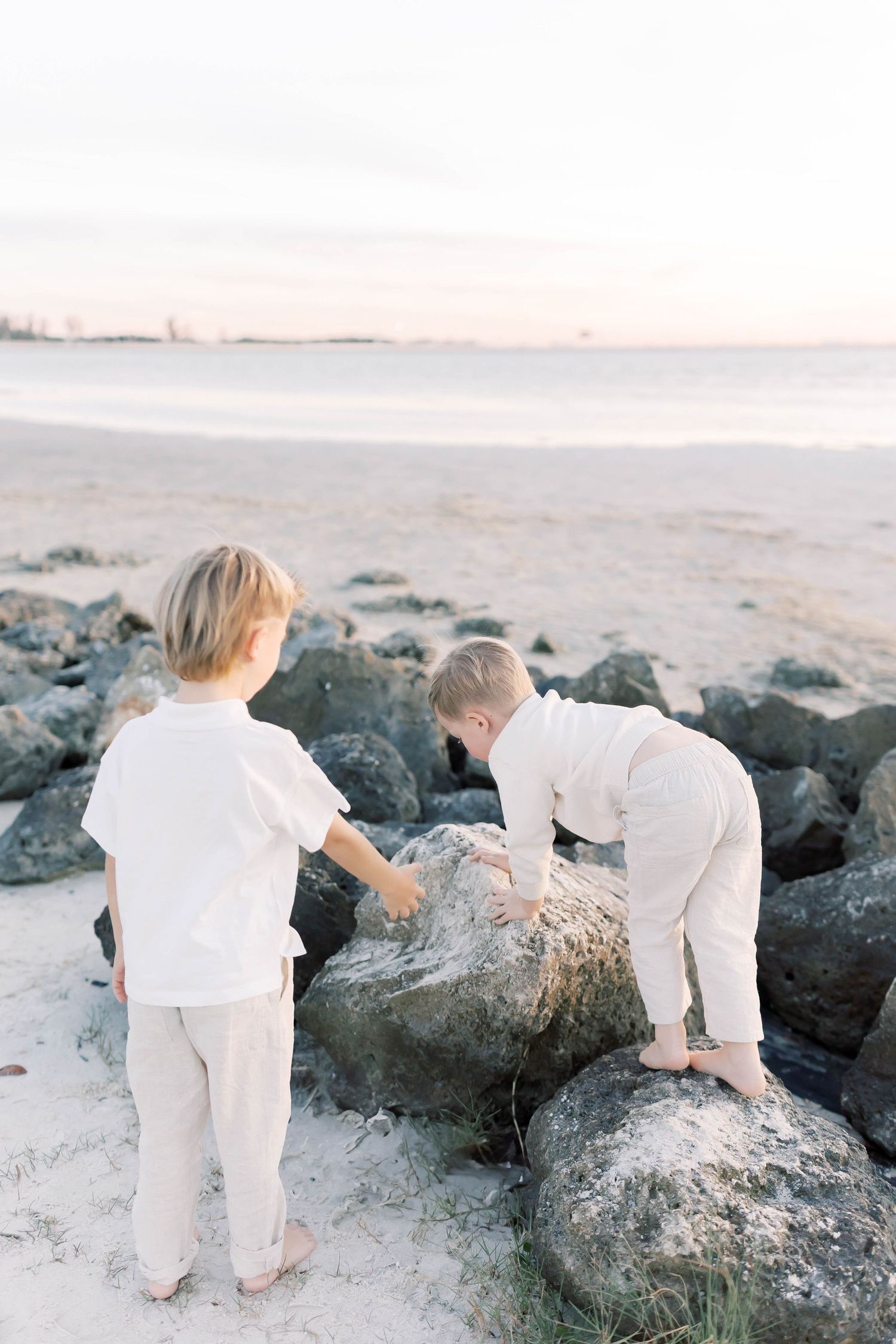 Children in white outfits explore large rocks on sandy beach at sunset.