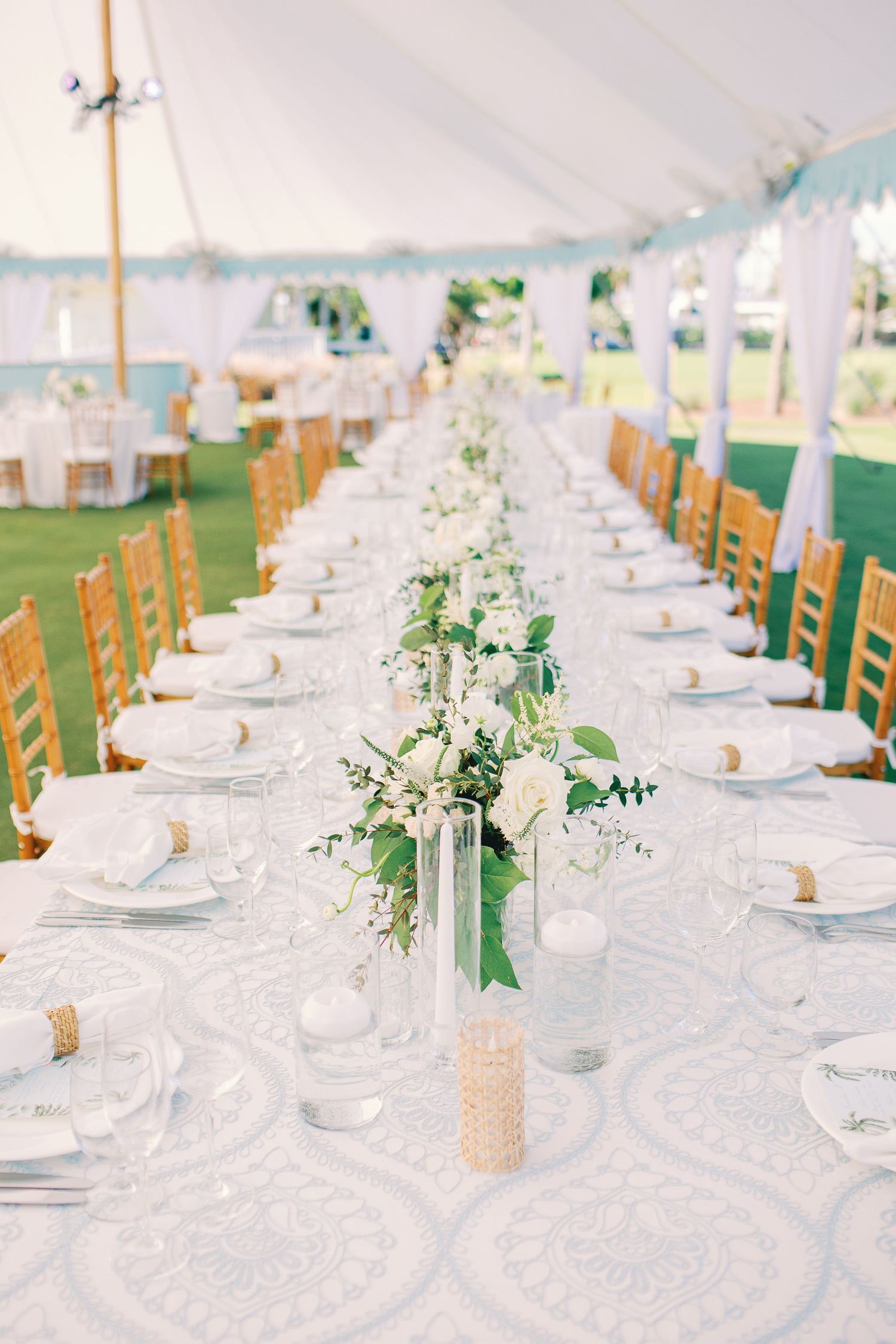 Elegant outdoor wedding reception under a white tent with long tables, white lace linens, gold chairs, and floral centerpieces.