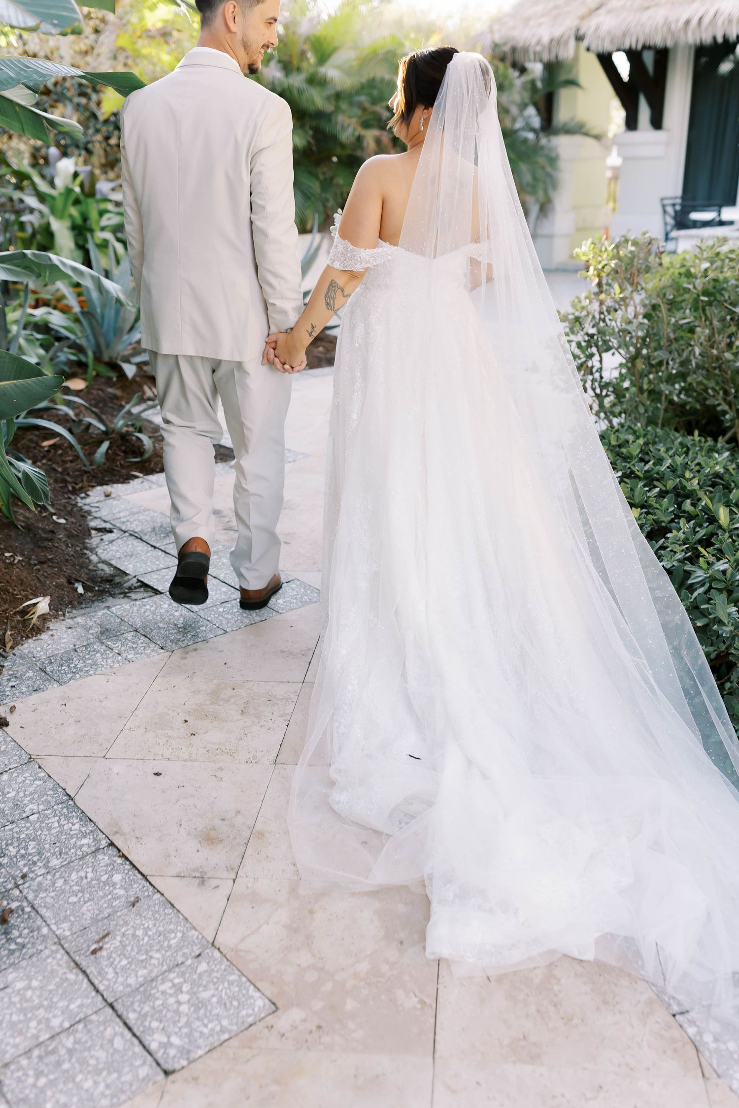 Bride and groom walk hand in hand along a garden pathway, bride's long veil trailing behind.