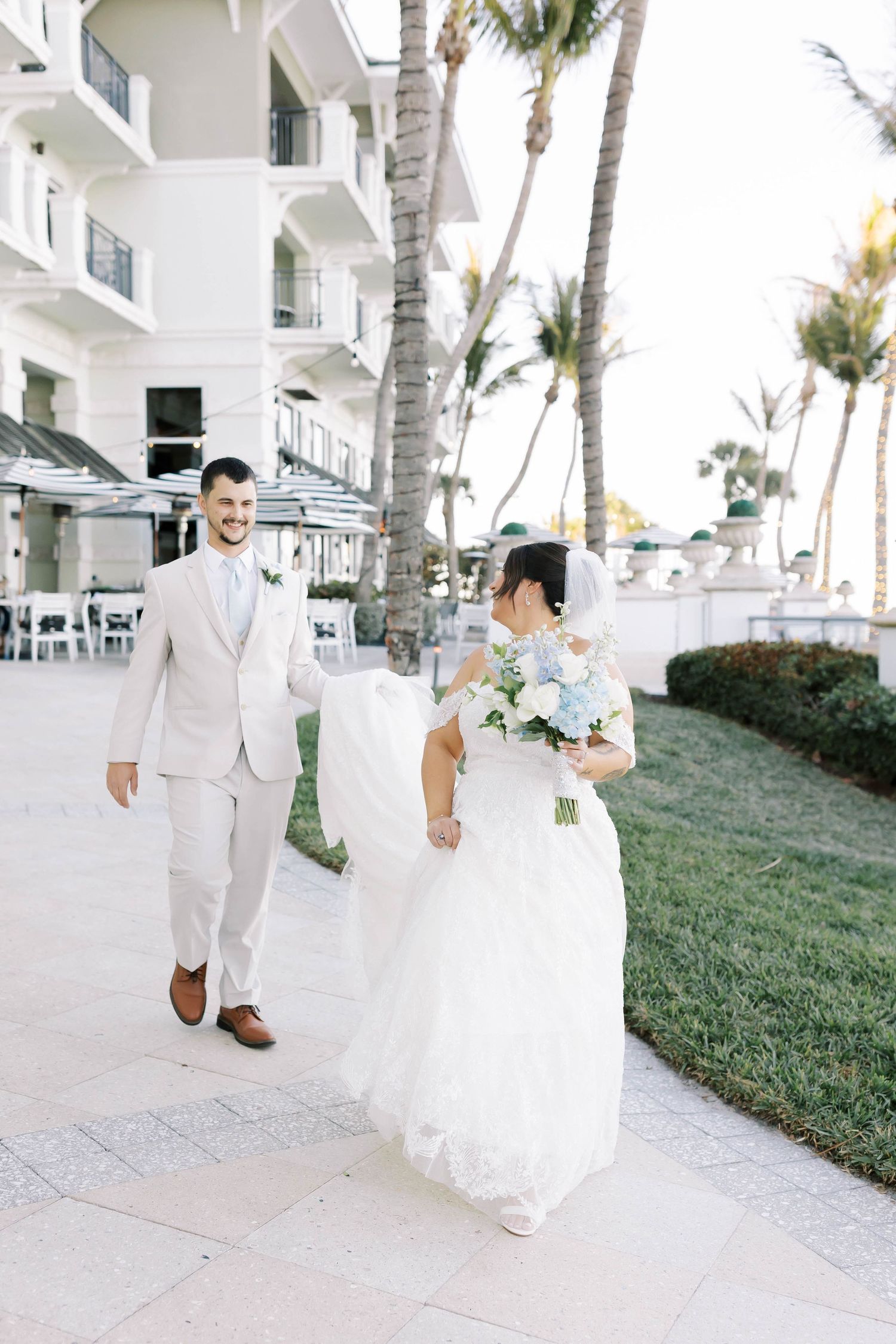 Bride and groom walk hand-in-hand along a tropical resort pathway, bride holding a blue and white bouquet.