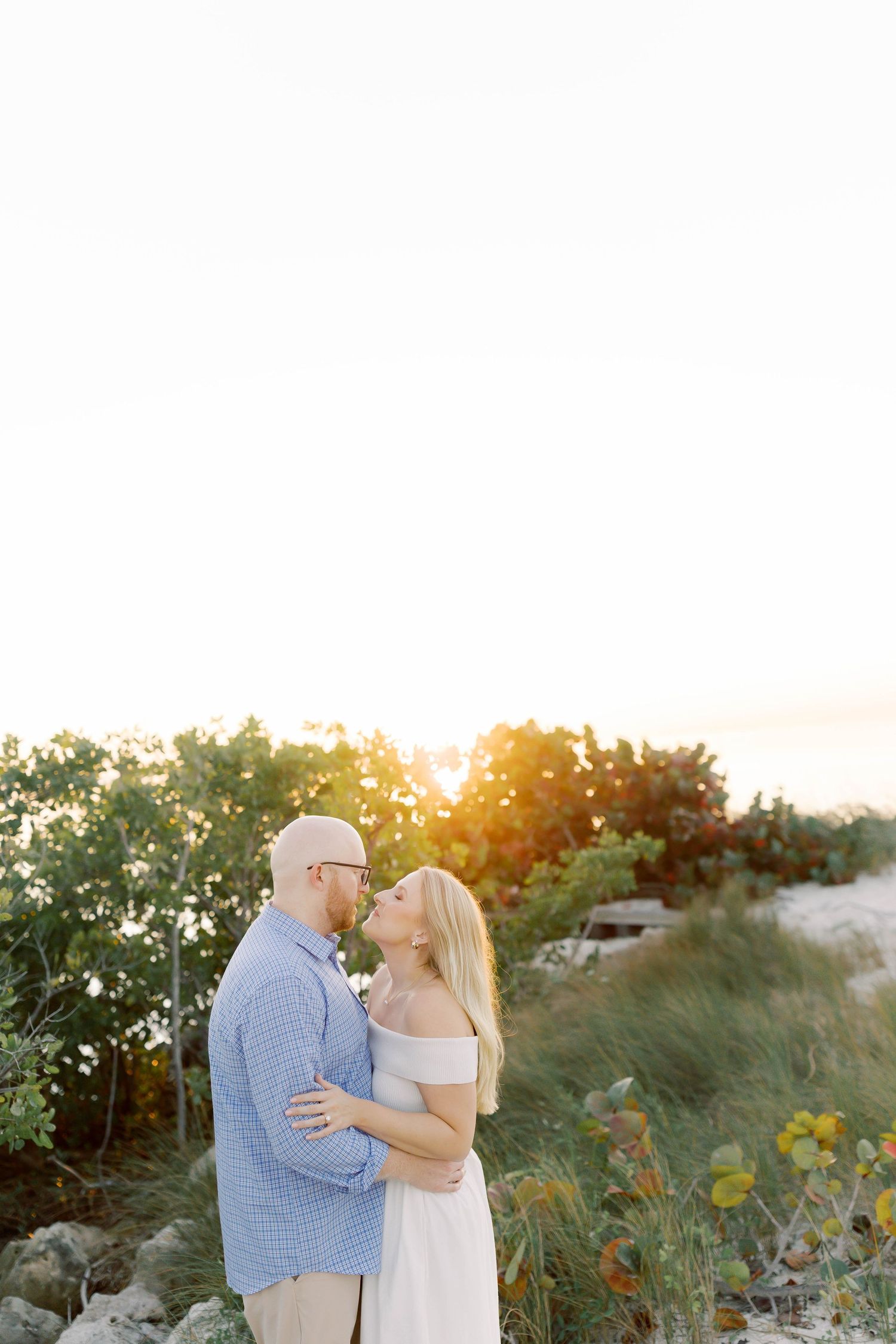 Couple embraces among coastal dunes and vegetation during golden hour.