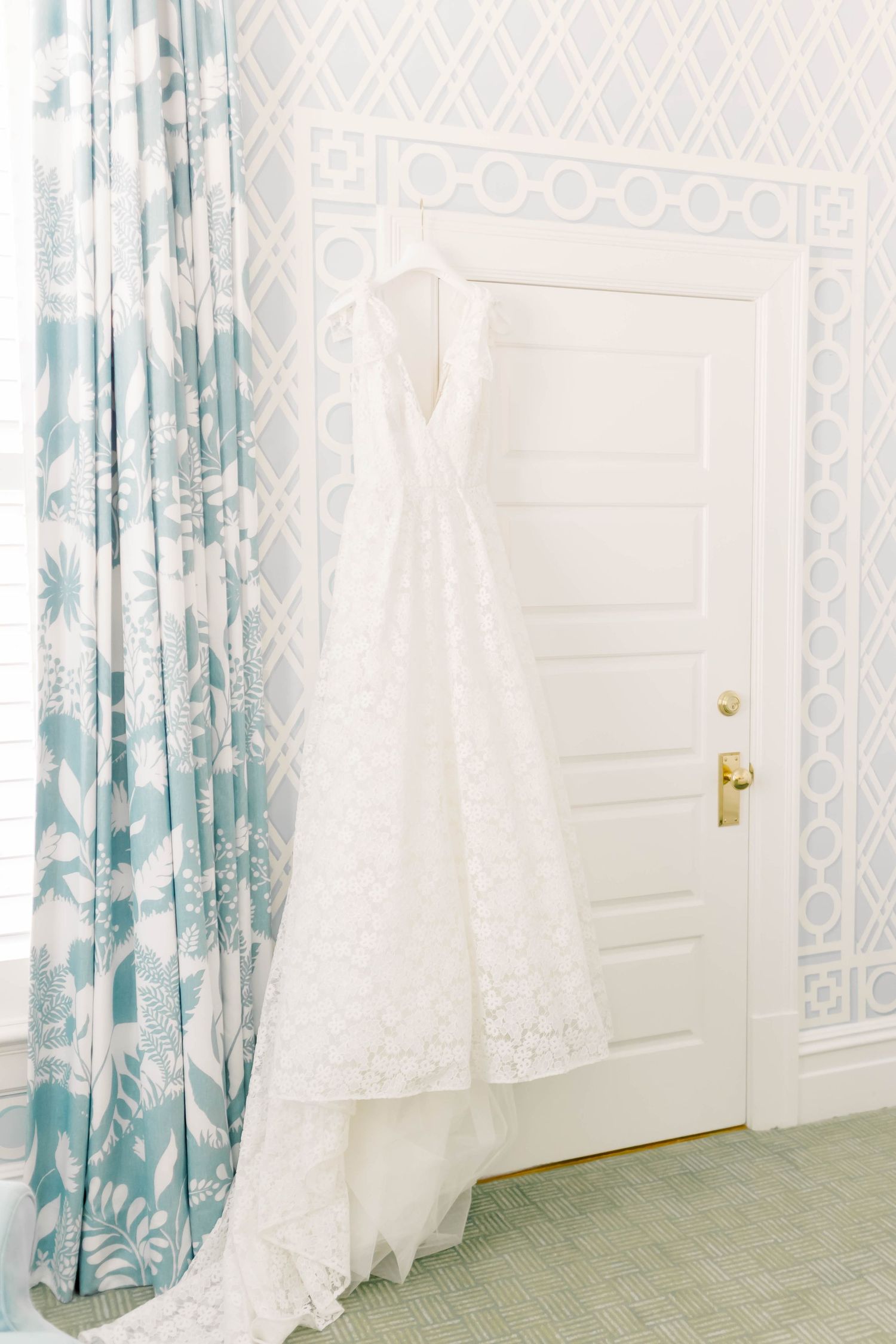 White wedding dress hanging in a room with blue floral curtains and geometric wallpaper.