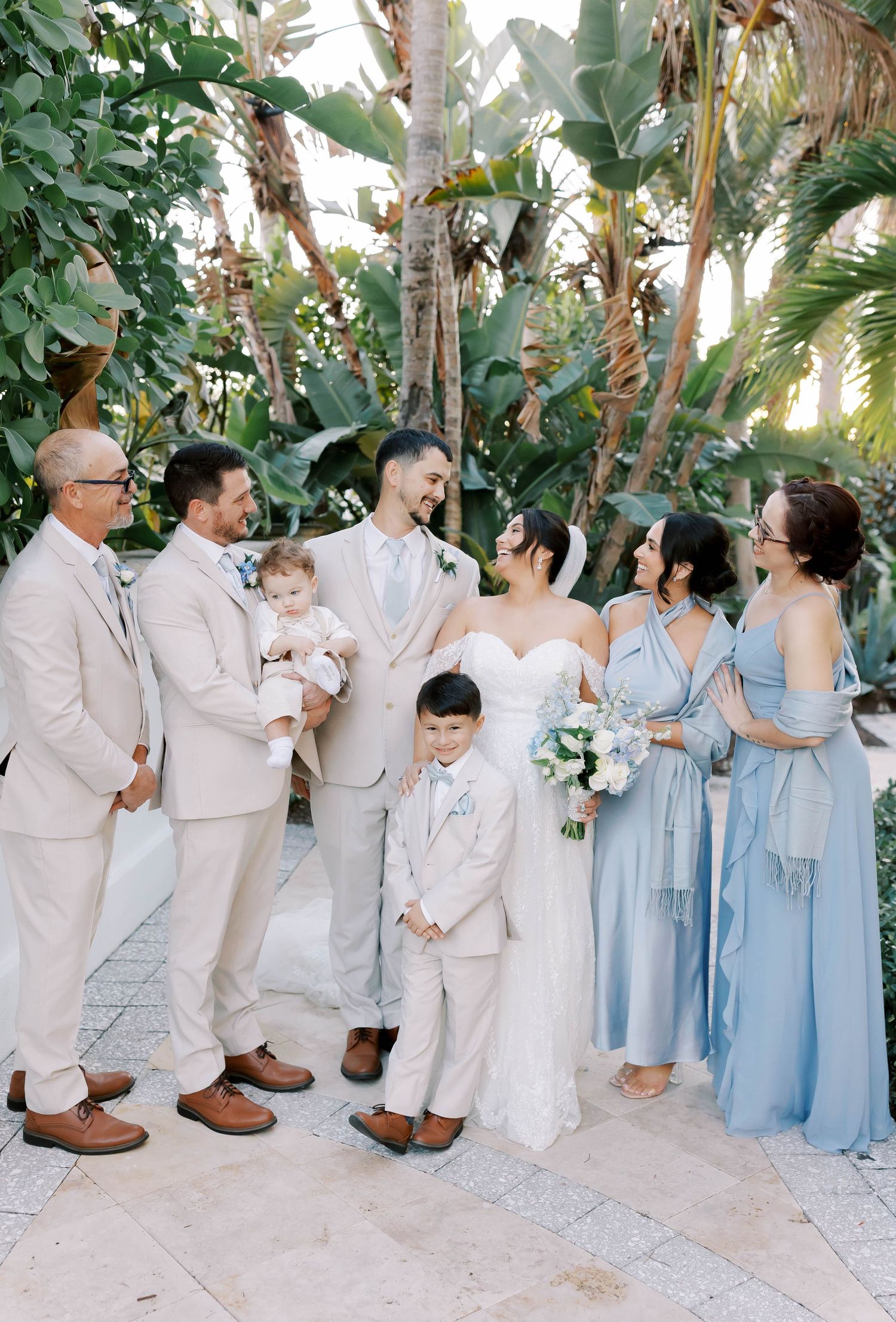Wedding party poses with bride and groom surrounded by tropical greenery in soft blue and beige attire.
