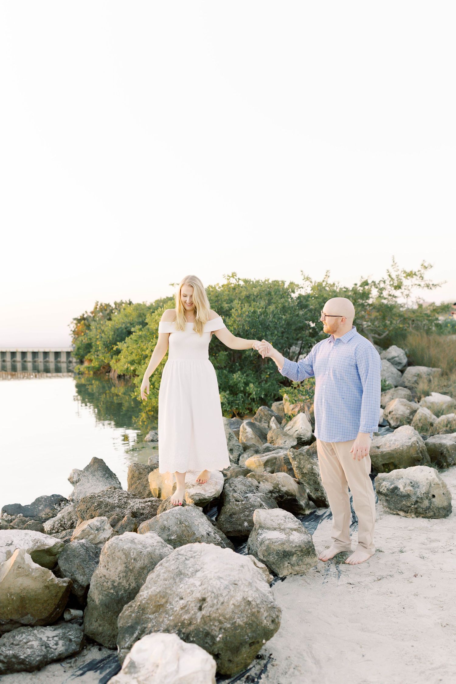 Couple holding hands on rocky shoreline, woman in white dress, man in blue shirt, with water and greenery behind them.