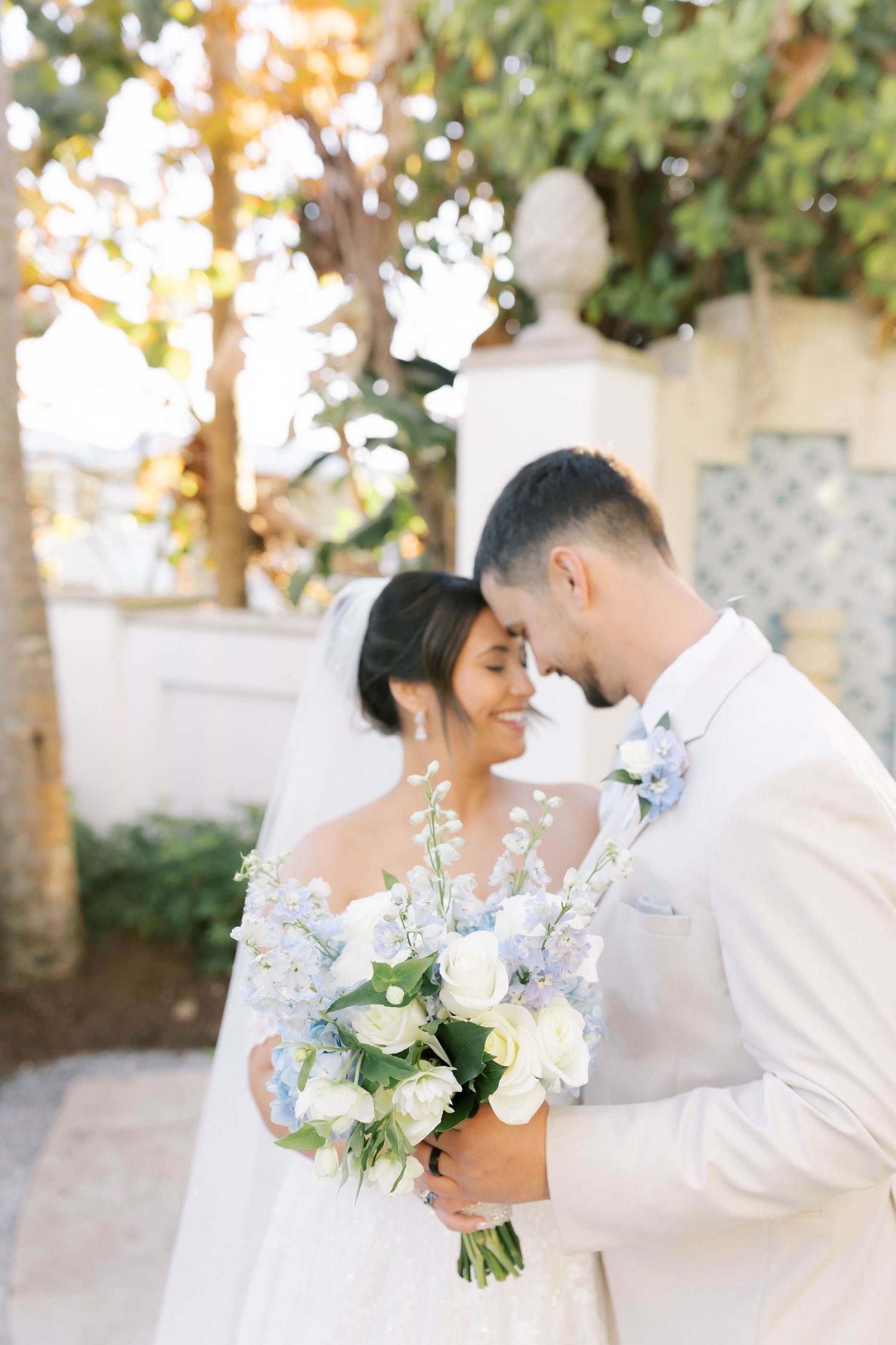 Bride and groom in white attire share a romantic moment holding a blue and white floral bouquet outdoors.