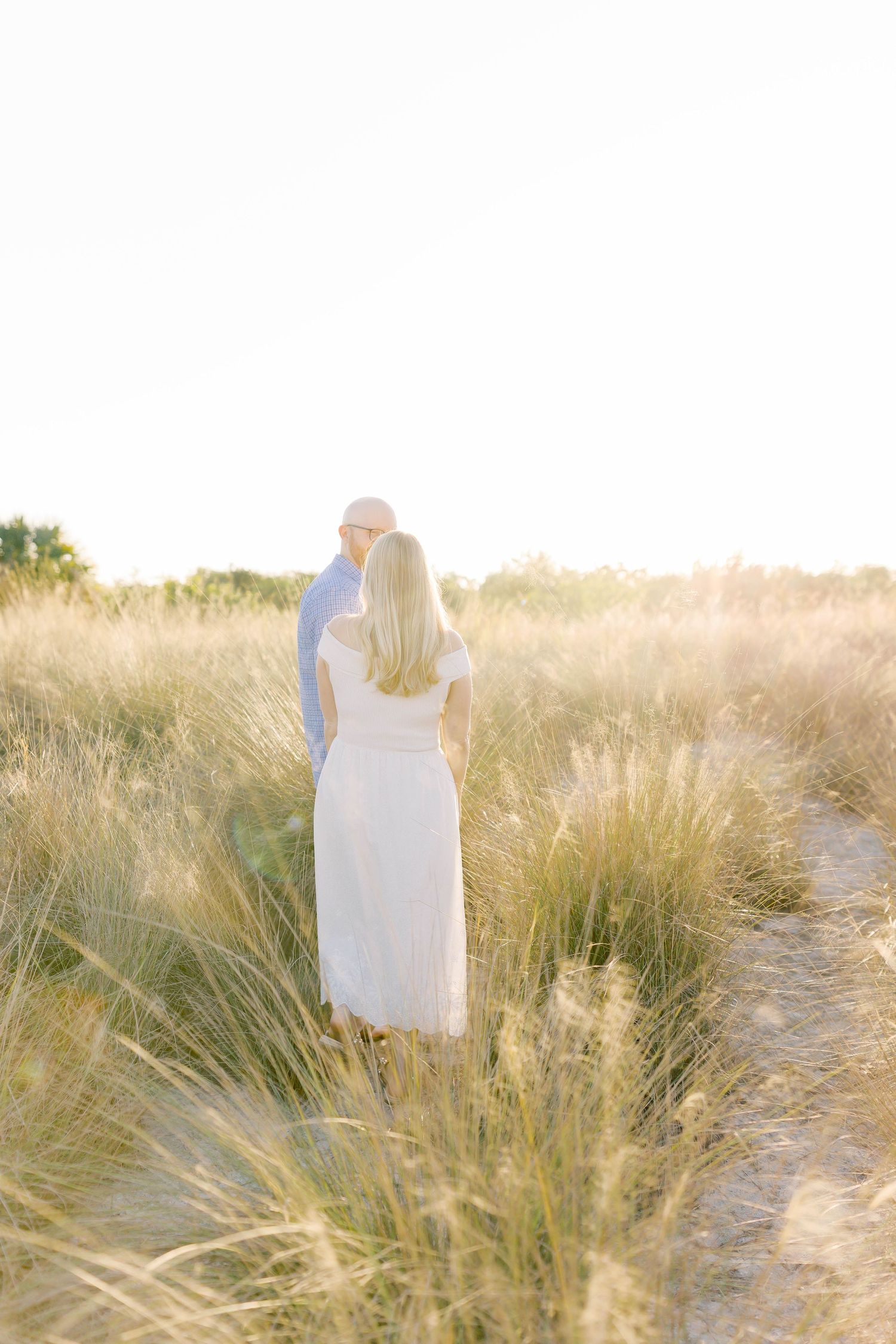 Couple stands together in sunlit grass field during golden hour photography session.