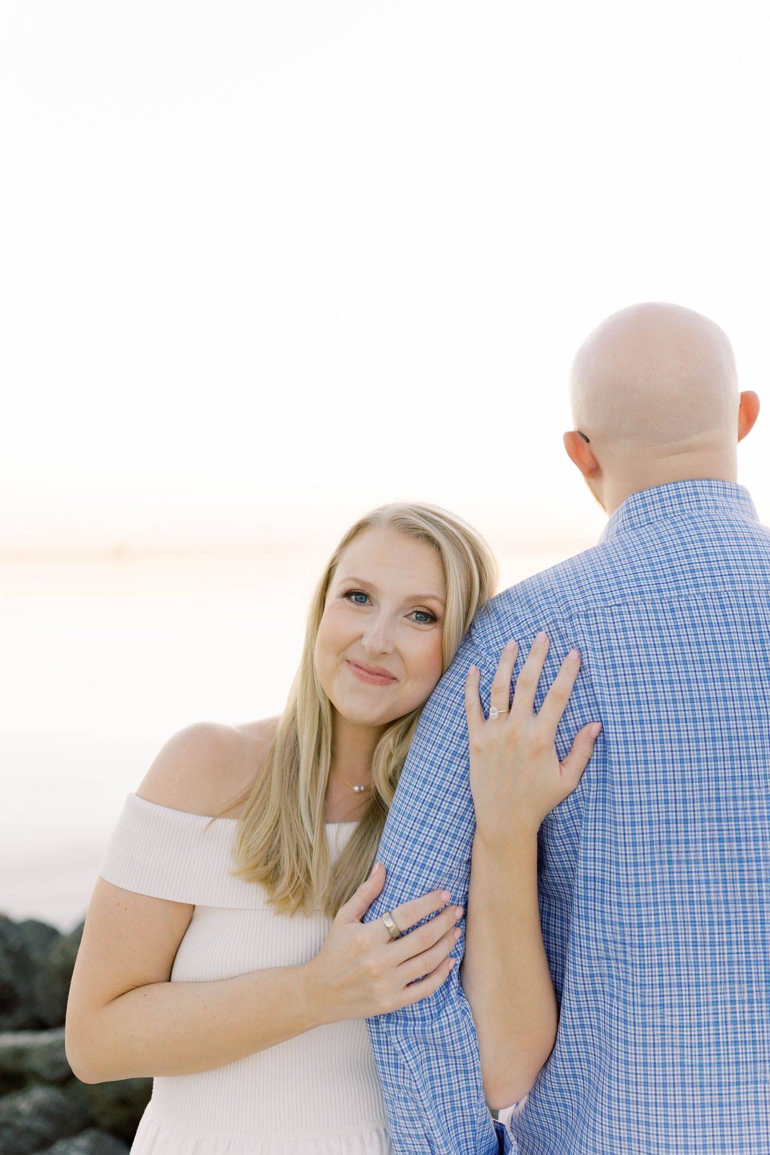 A romantic couple embraces during a sunset photoshoot, with the woman wearing a white dress and man in blue plaid.
