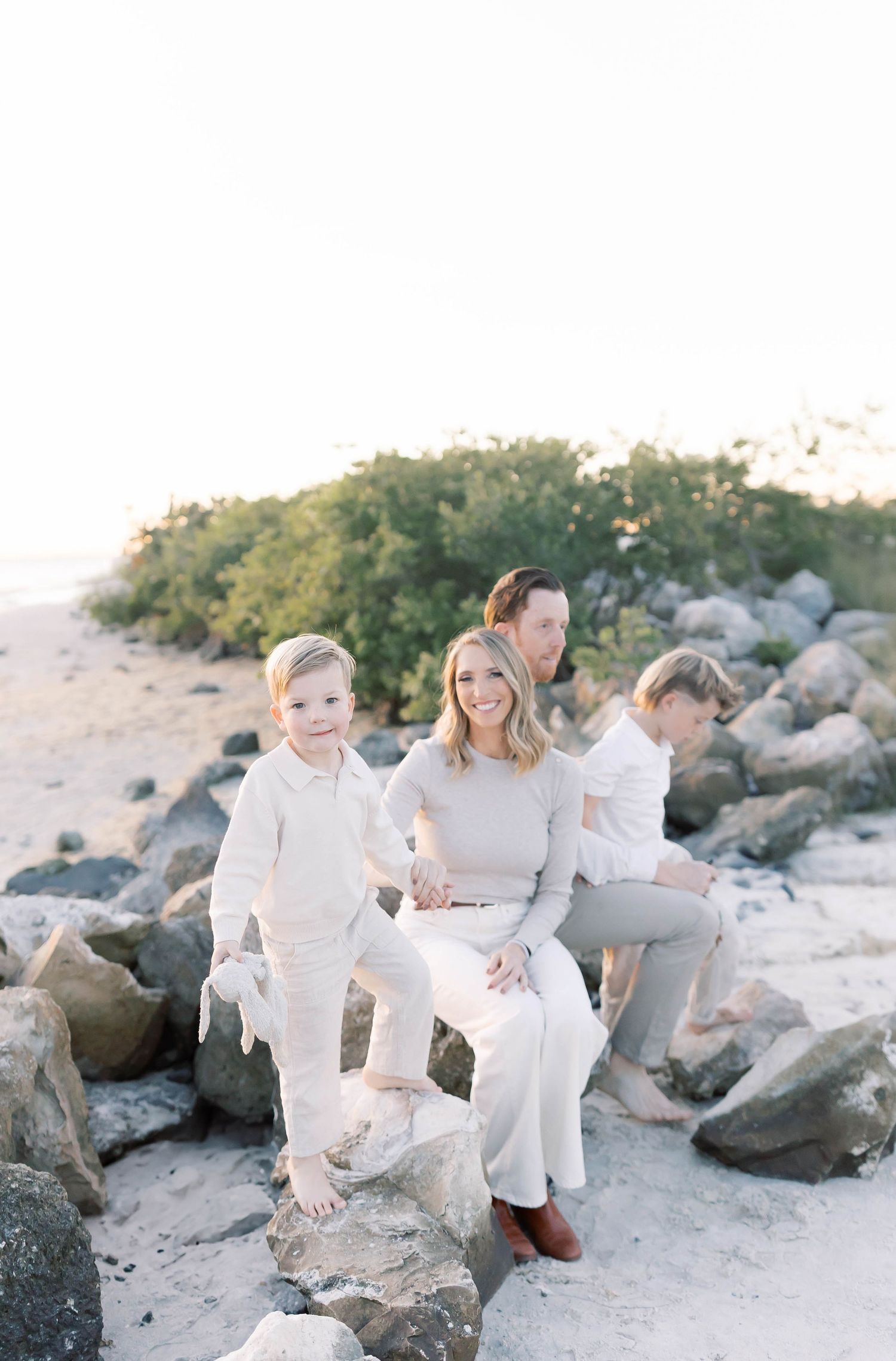 Family wearing white outfits sits together on rocky beach during sunset photoshoot.