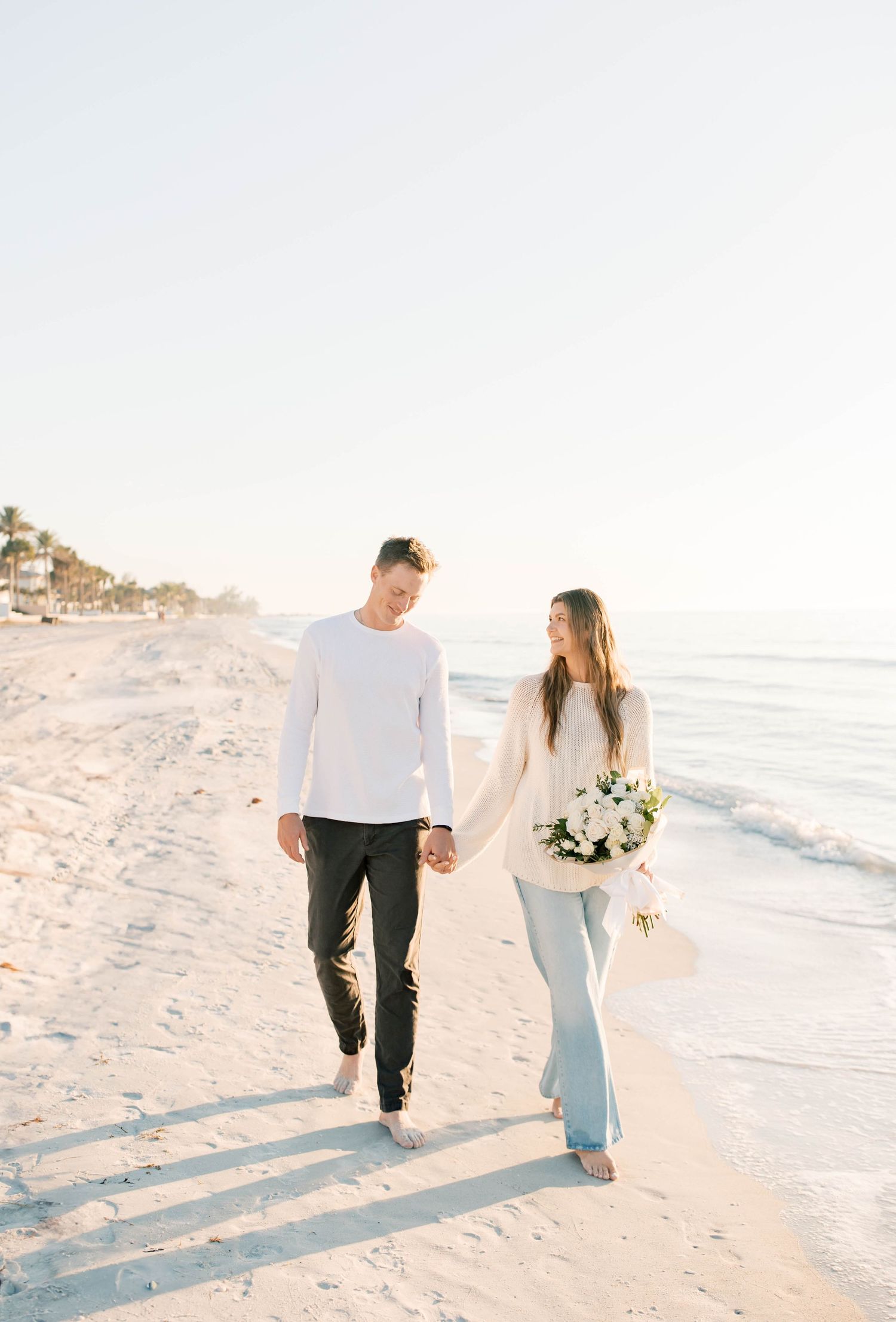 A couple walks along white sandy beach during sunset, one holding a bouquet of flowers.