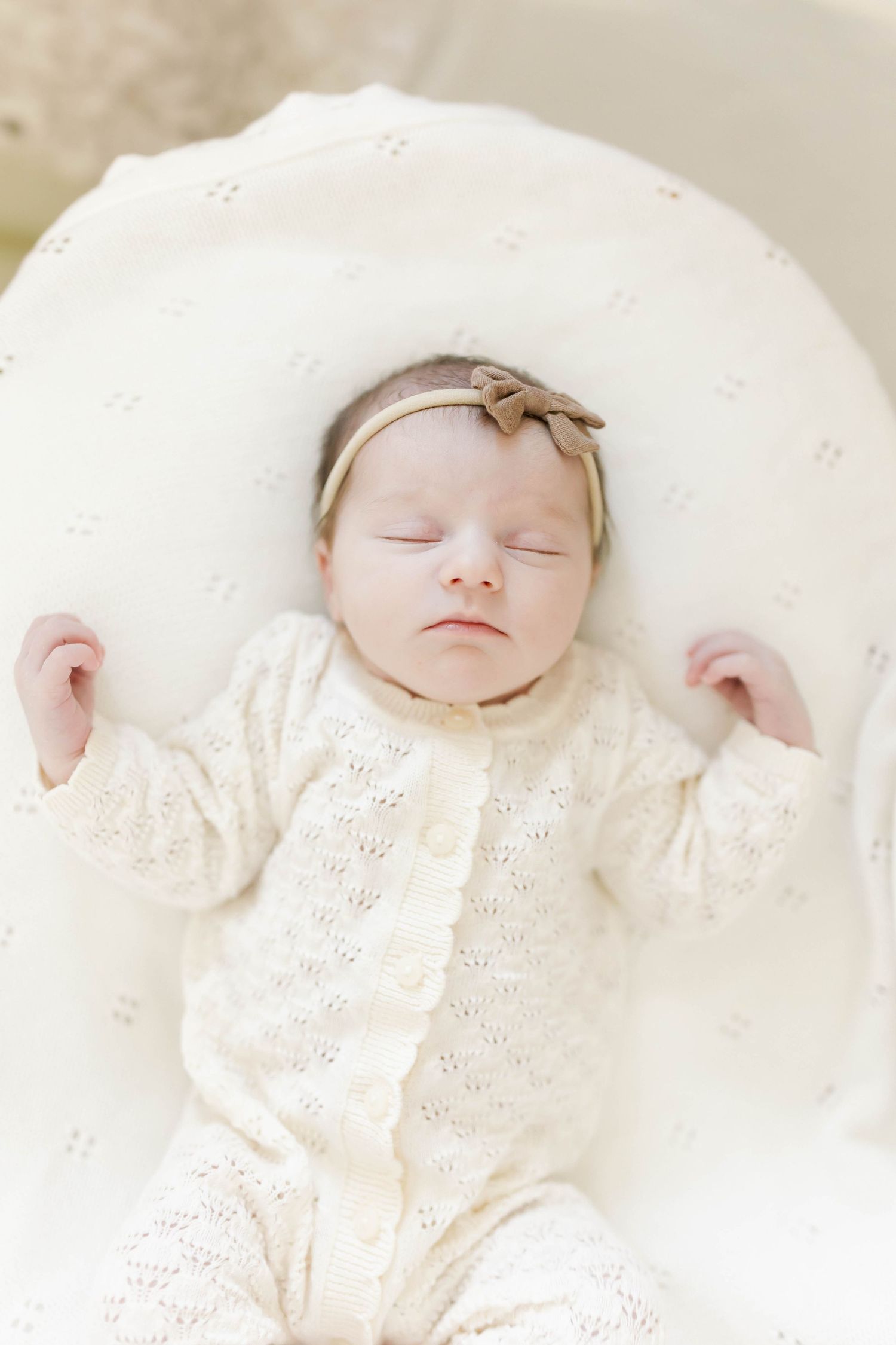 Sleeping newborn wearing delicate white lace dress rests peacefully on soft white bedding.