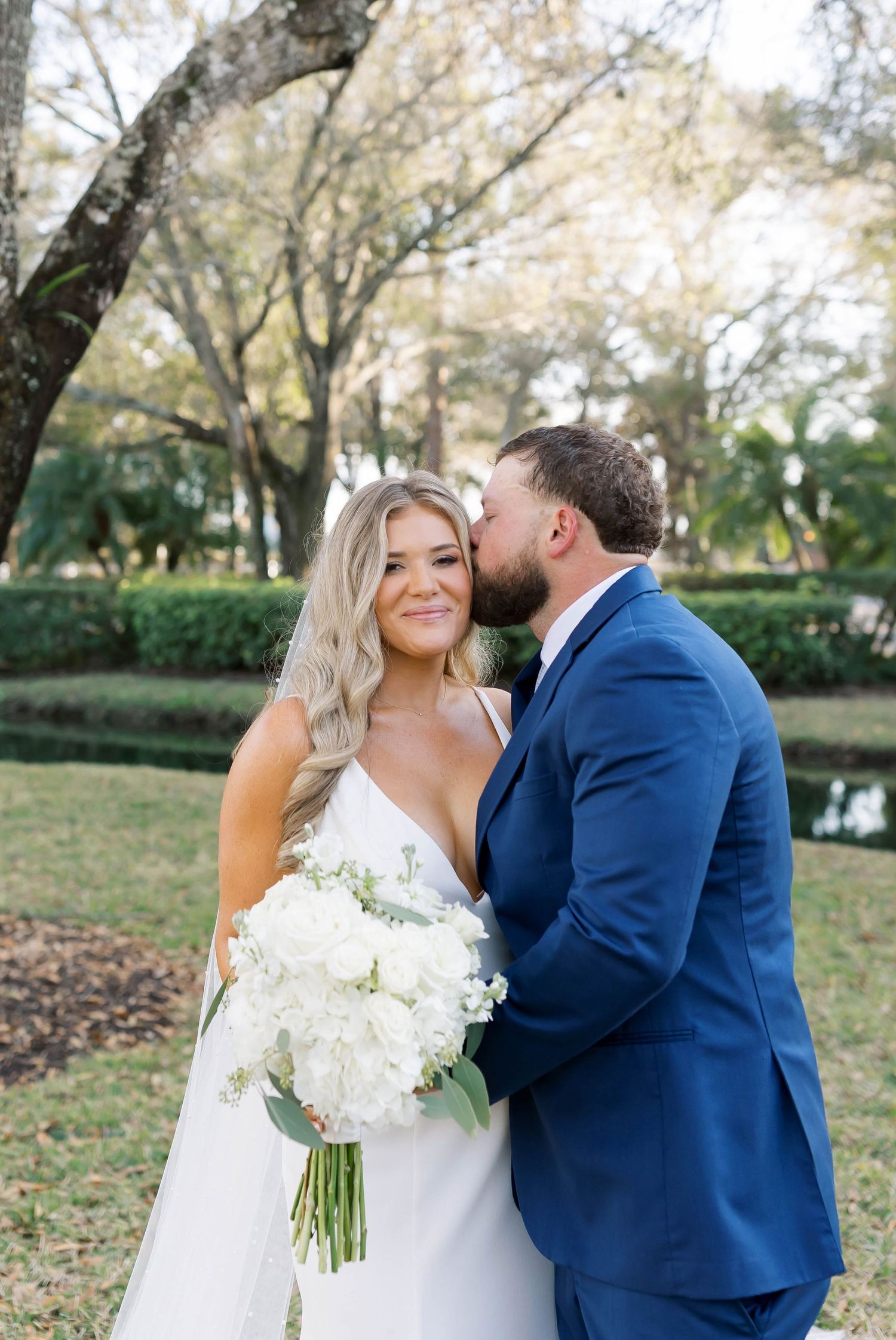 Bride holding white bouquet smiles as groom in blue suit kisses her cheek outdoors among lush green trees.