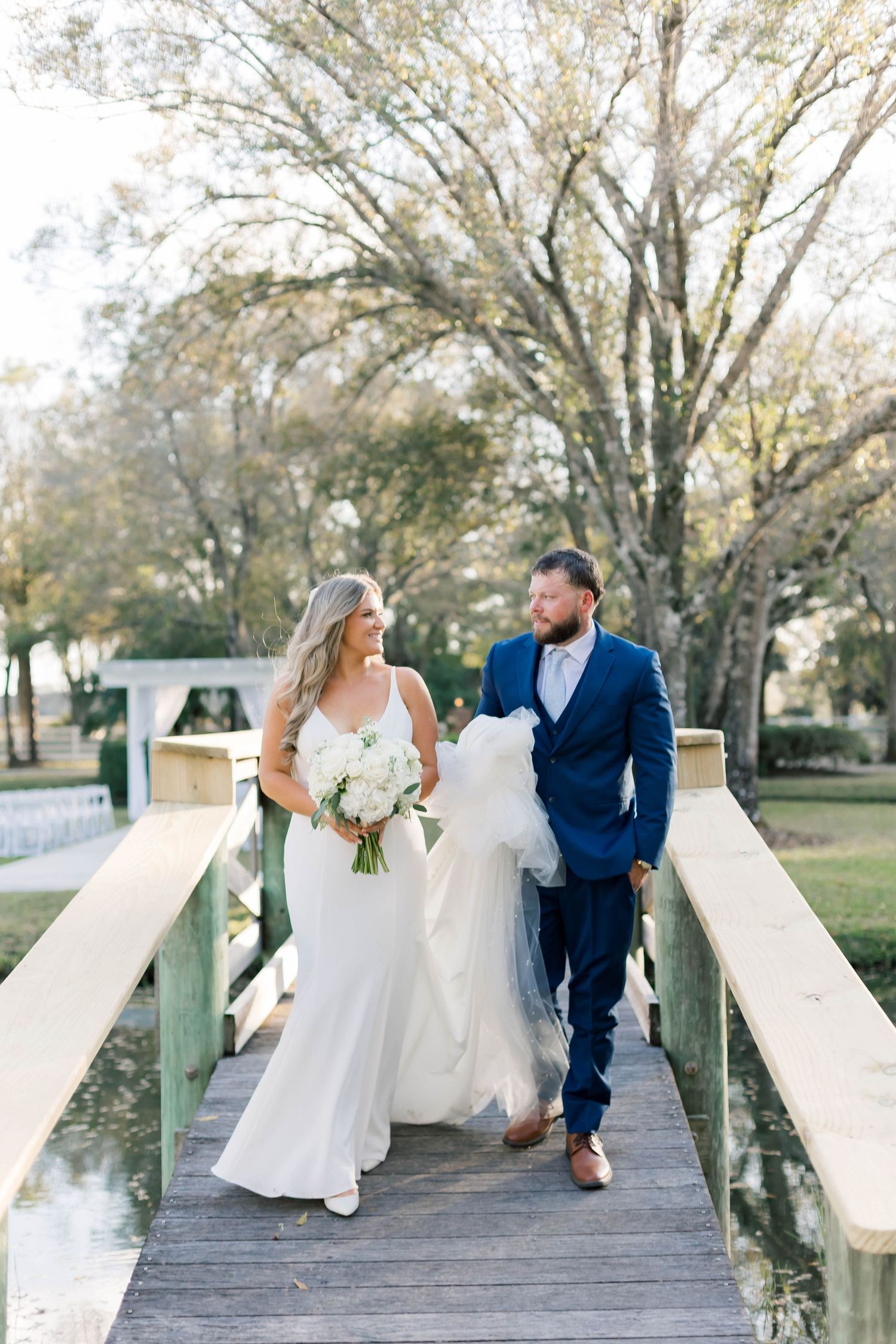 Newlyweds walking together on a wooden bridge holding a white bouquet in an outdoor garden setting.