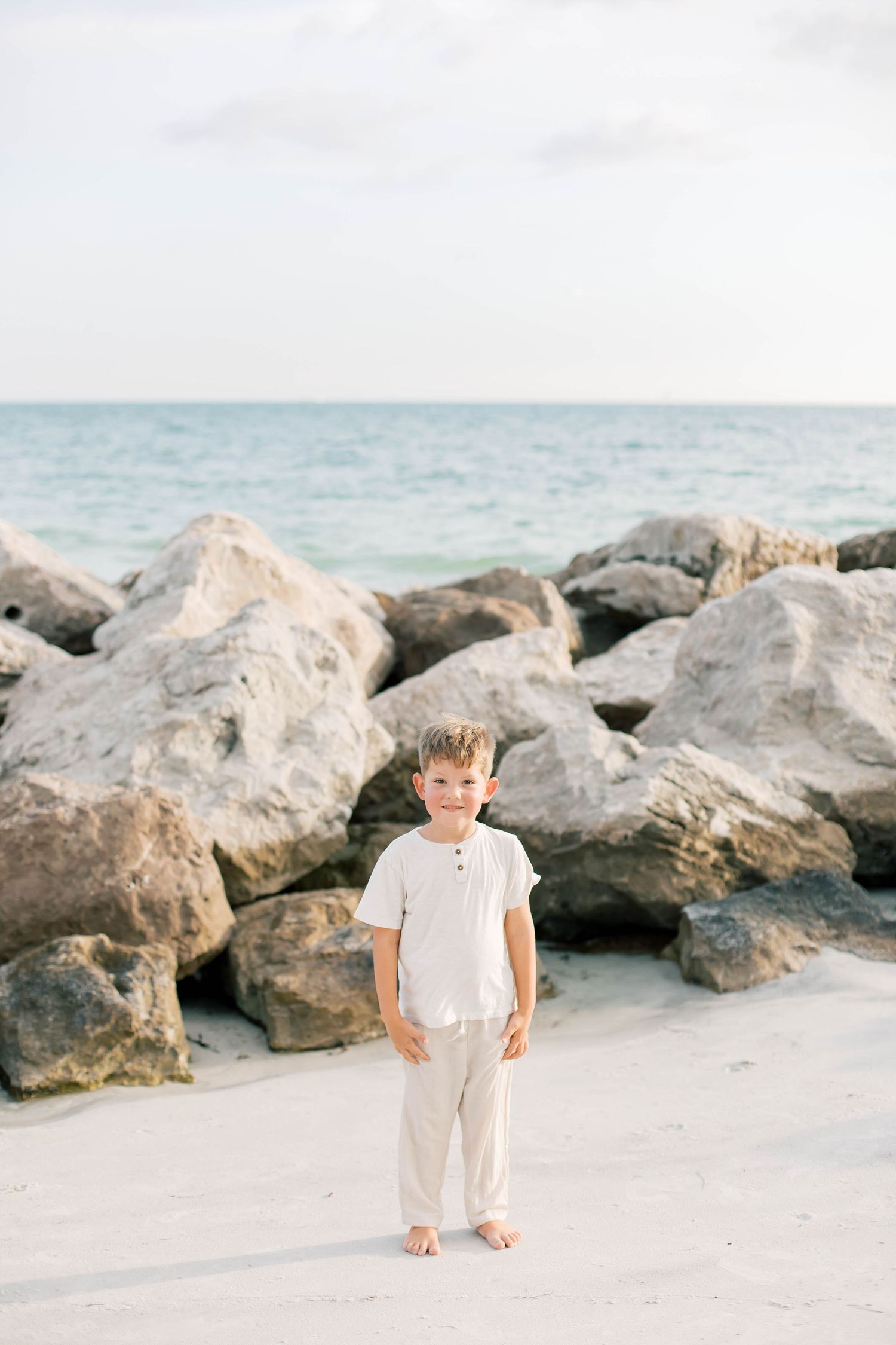 A toddler in light colored clothing stands on a beach near large rocky formations.