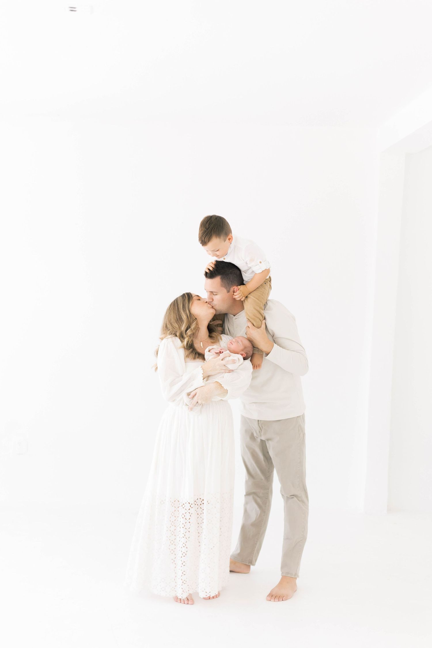 Family portrait session with parents and newborn in white studio setting.