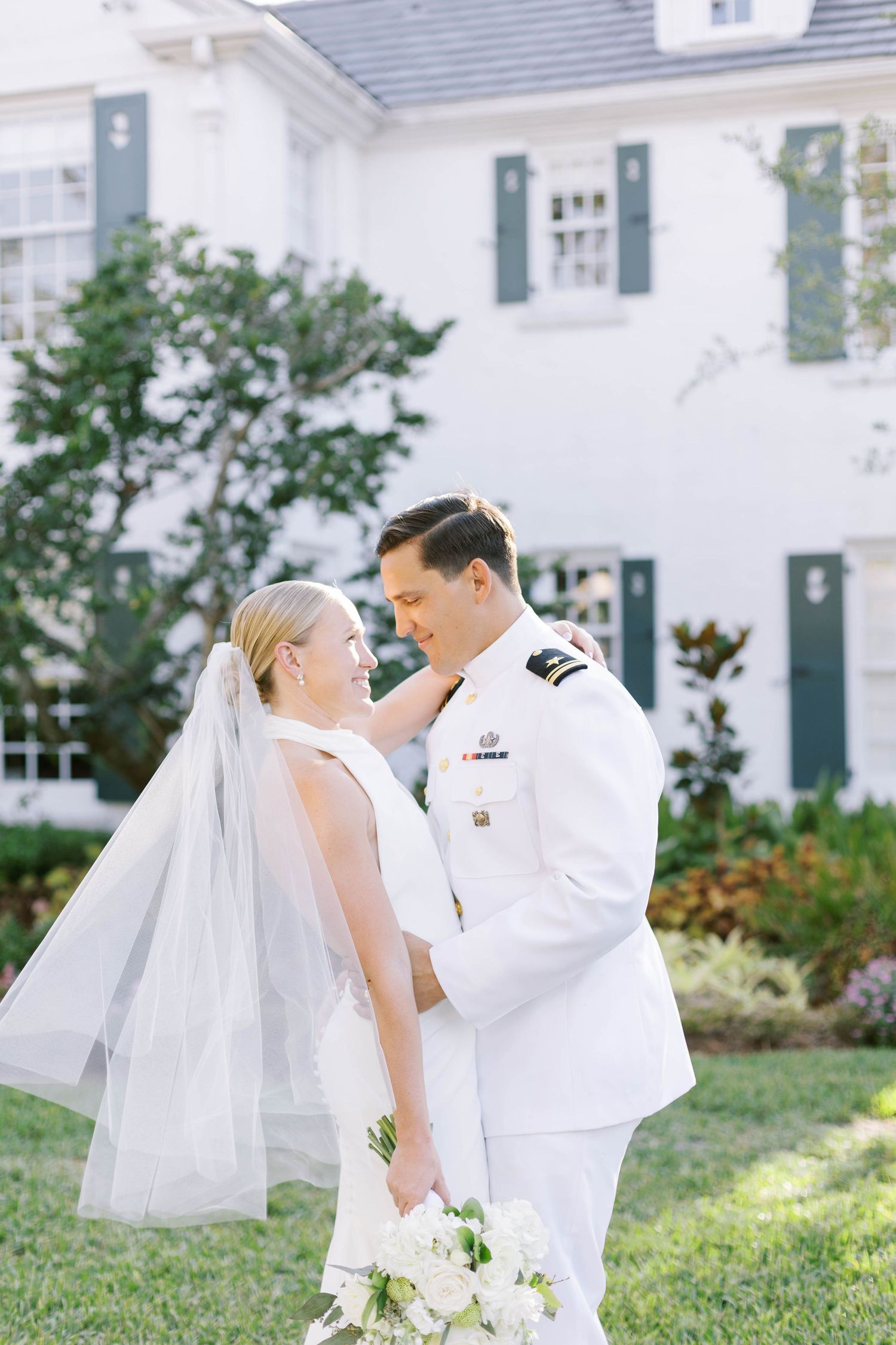 Bride and groom in military dress whites share a romantic moment outdoors in front of an elegant white colonial home.