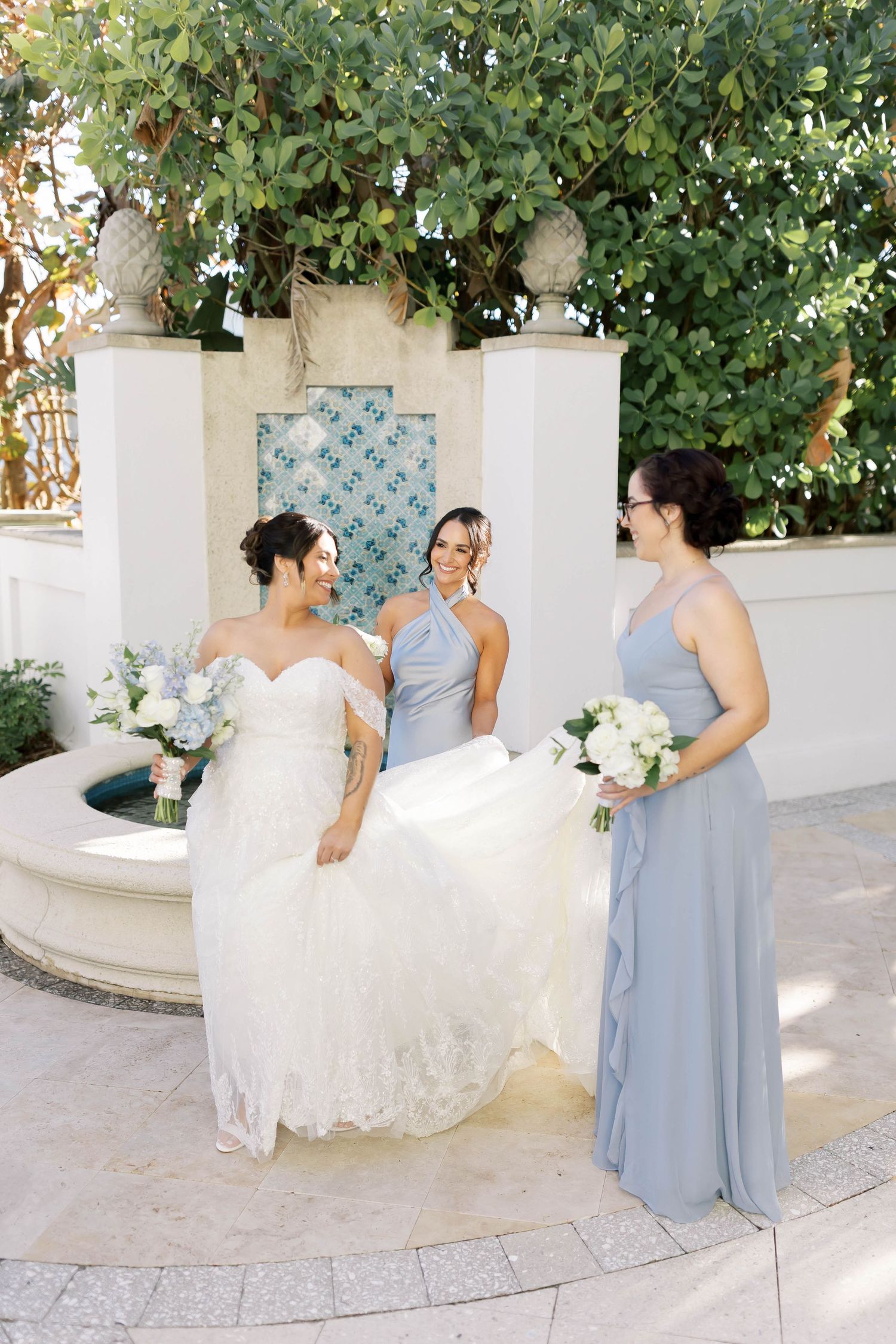 Bride in lace off-shoulder gown laughs with two bridesmaids in dusty blue dresses near fountain.
