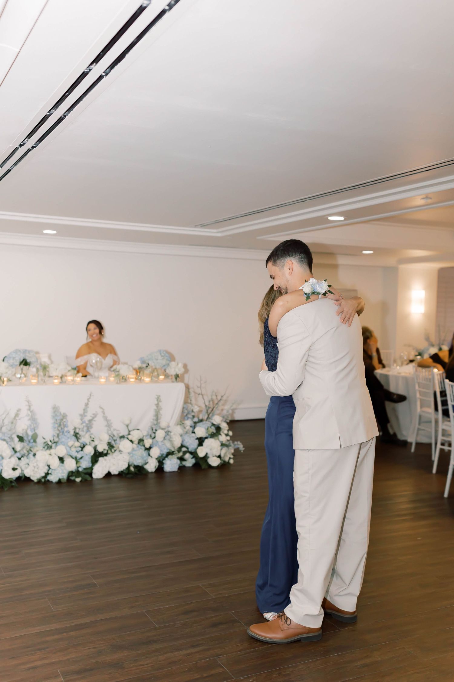 Groom in beige suit dances with mother in navy lace gown before lush white floral tablescape.