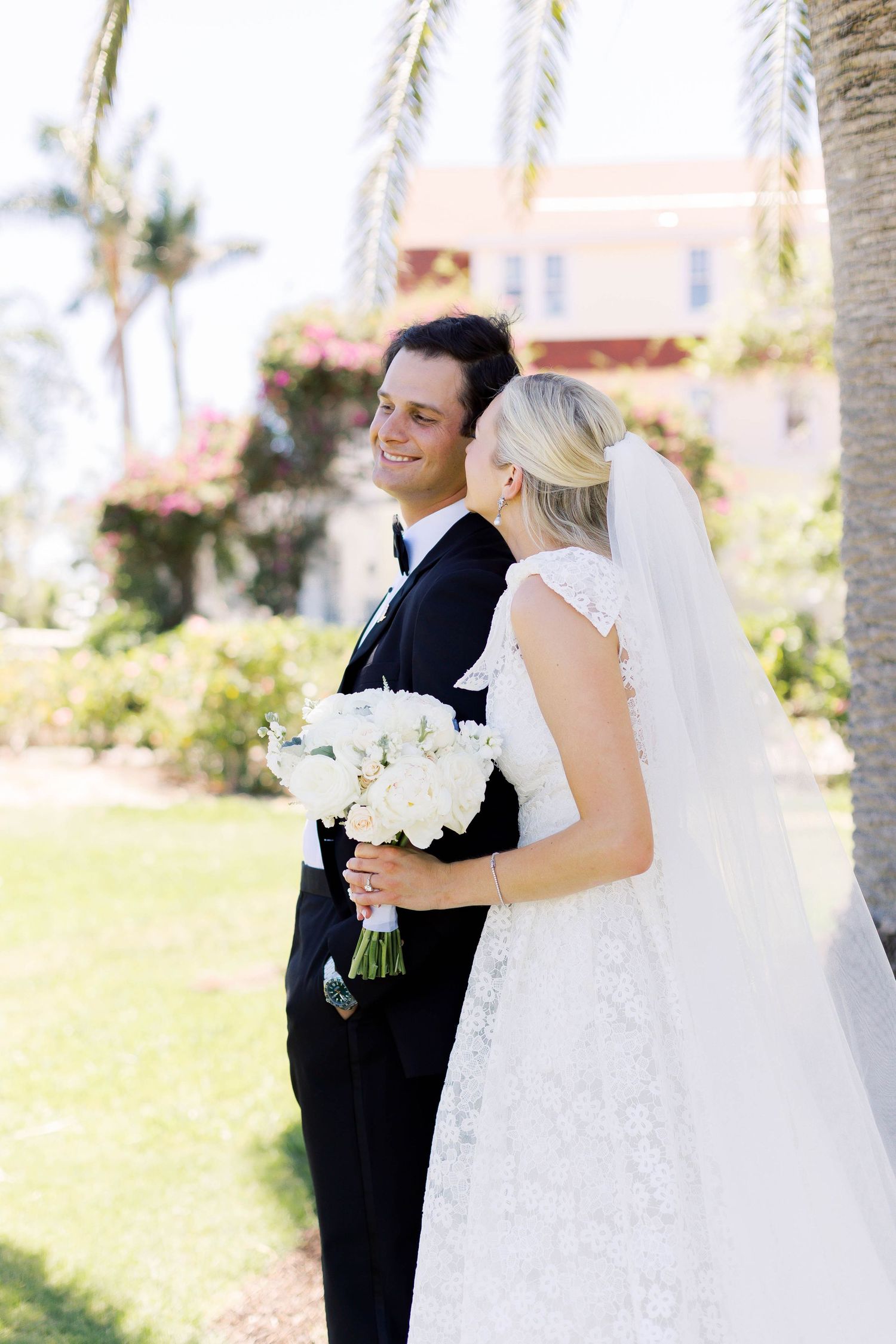 Smiling groom in black tuxedo with bride in lace wedding dress holding white bouquet outdoors near palm trees.