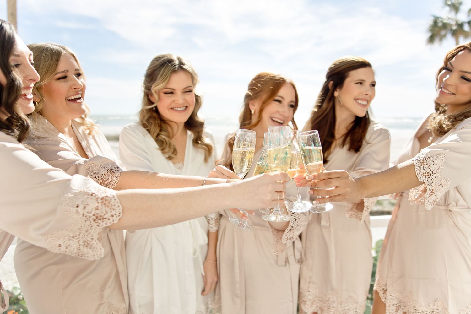 Women in blush robes toast with champagne glasses during a pre-wedding celebration.