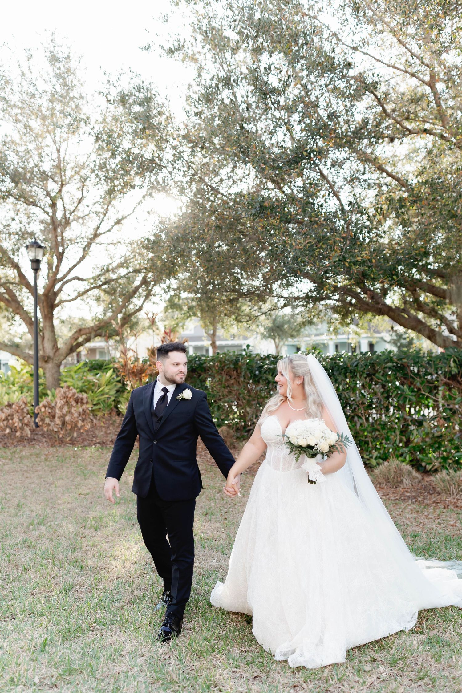 Bride and groom walk hand-in-hand outdoors, she in a white gown with veil, he in a black suit, under lush trees.