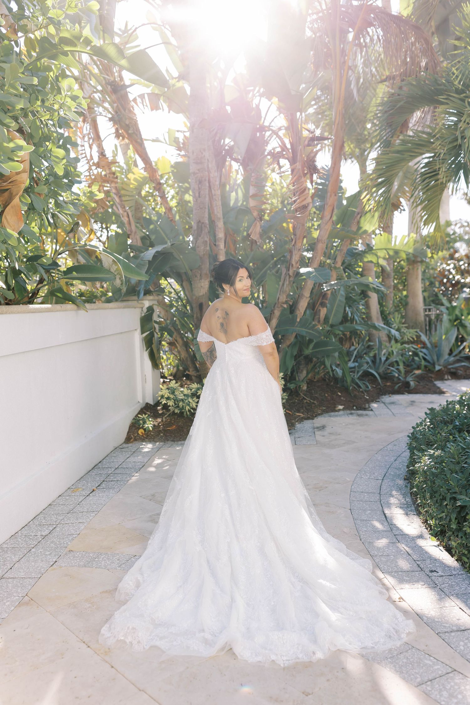Bride in off-shoulder lace wedding gown with long train stands on stone path surrounded by lush tropical gardens.
