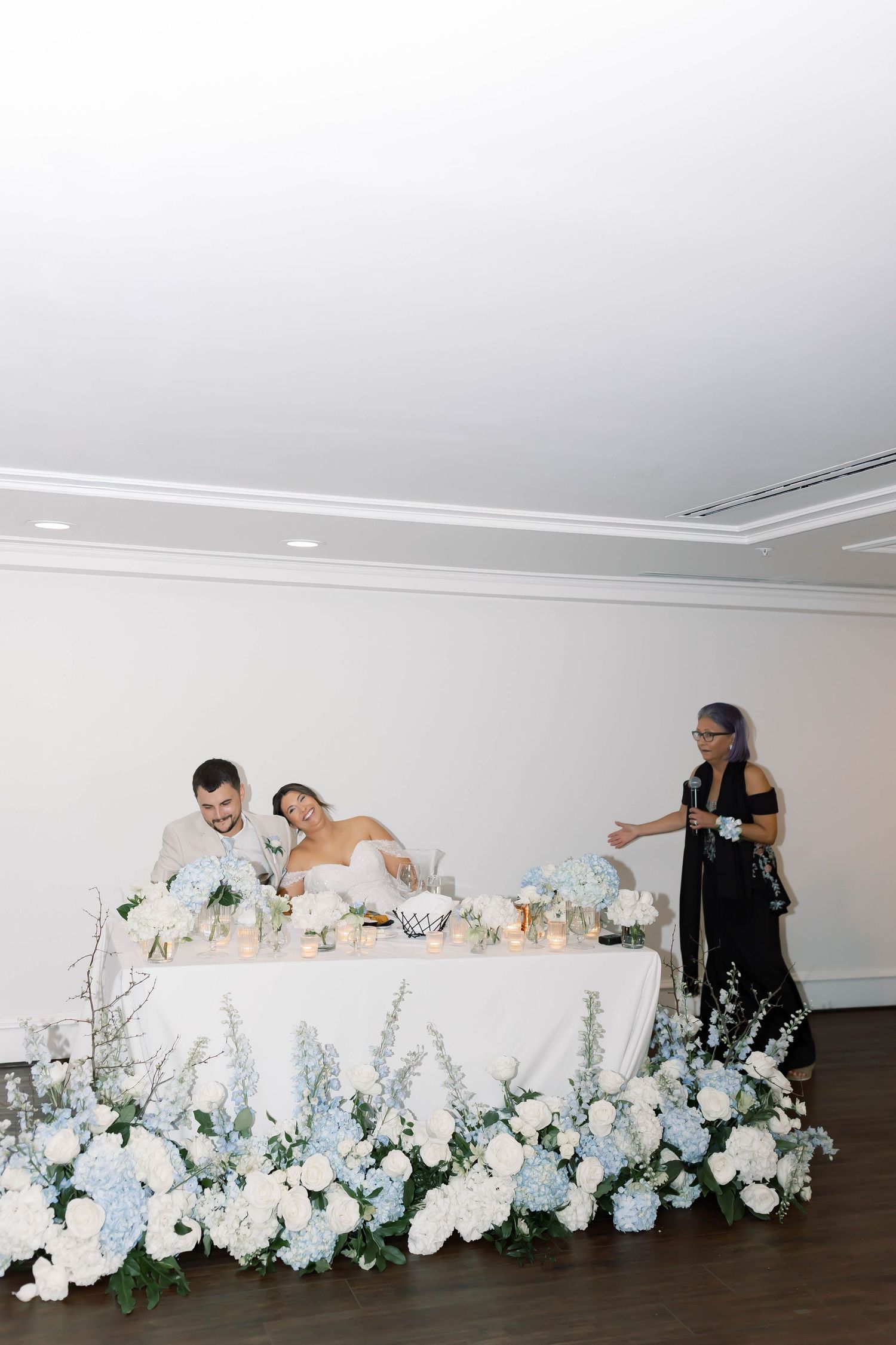 Bride and groom sit at a sweetheart table adorned with blue hydrangeas and white floral arrangements at reception.