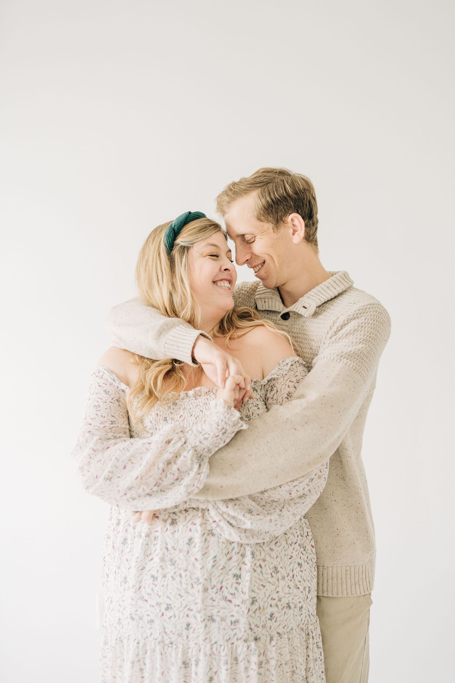 A smiling couple in neutral tones embracing closely, the woman in a floral dress with a green headband.