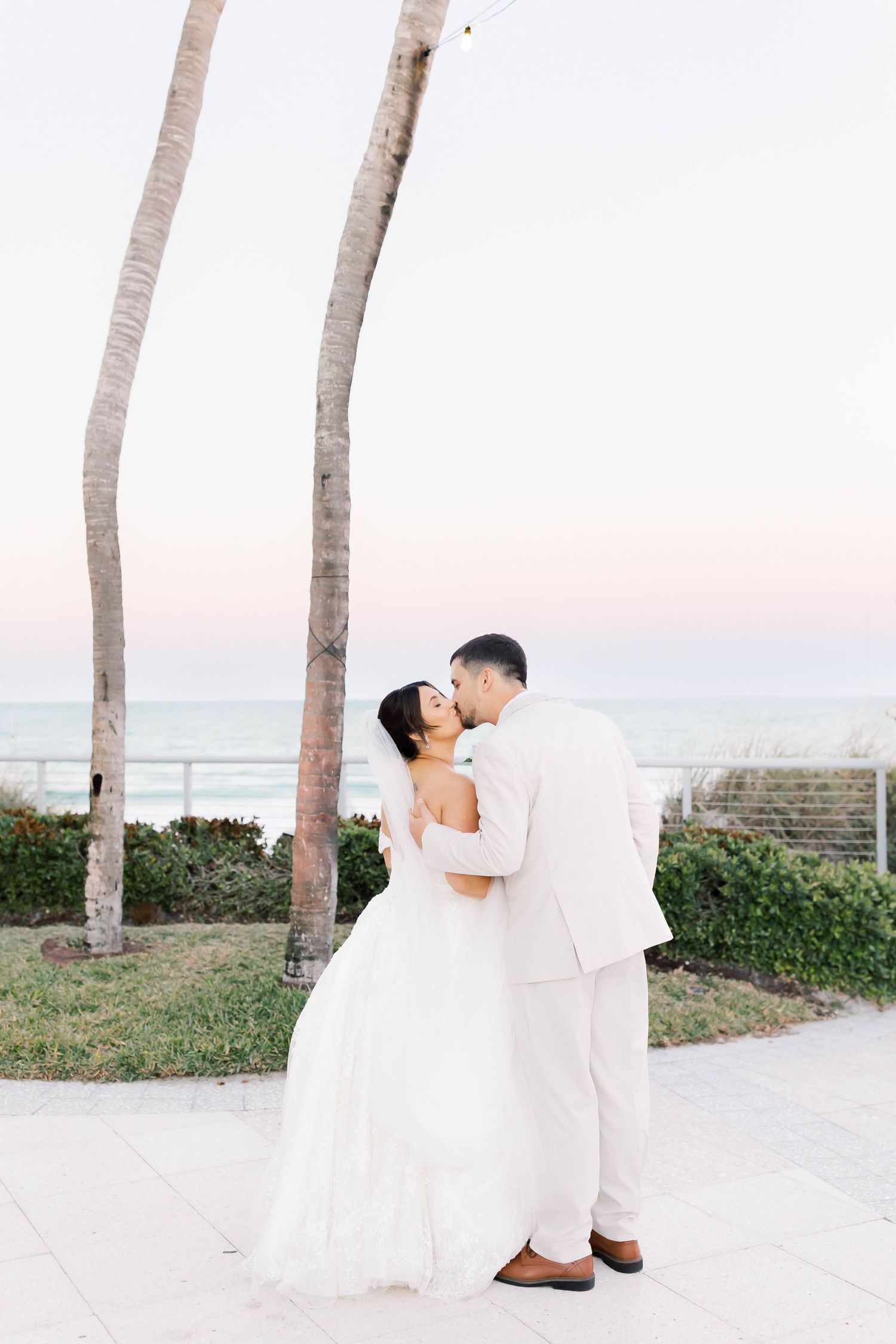 Bride and groom share a romantic kiss near palm trees with ocean views at sunset during their beach wedding.