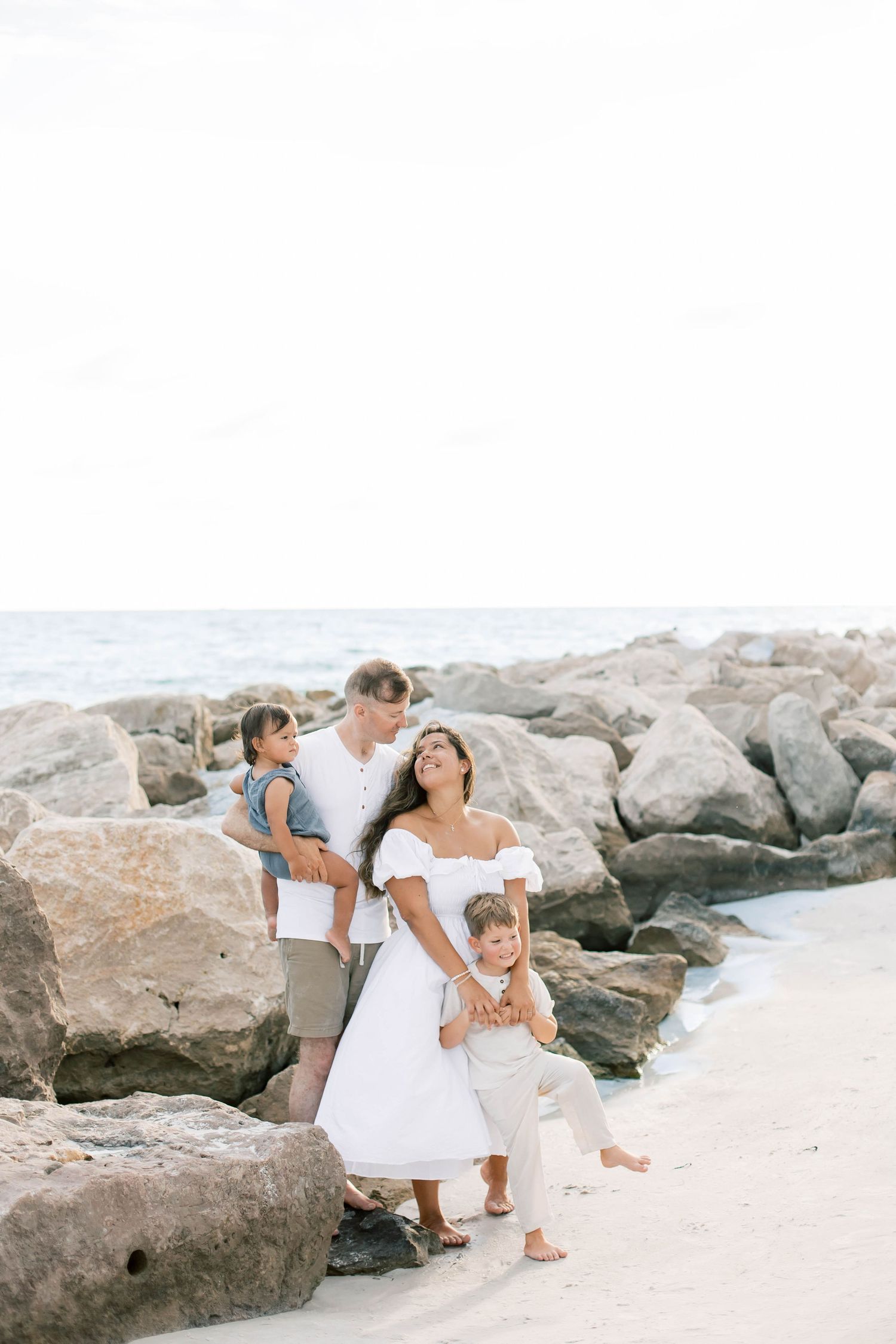 A family plays and embraces on a sandy beach with coastal rocks in the background.