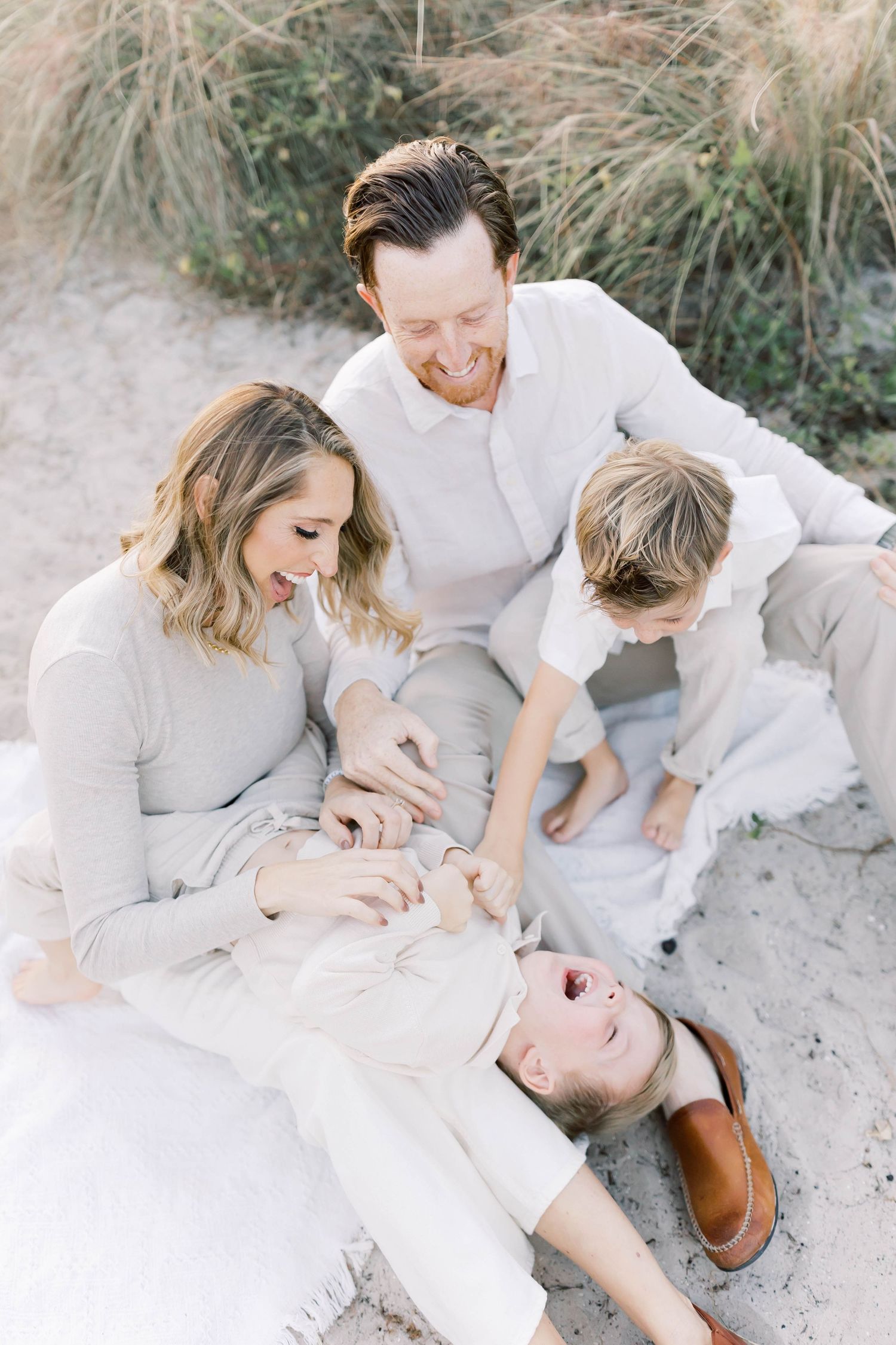 Family in light colored clothing plays and laughs together while lying on sandy beach during sunset photoshoot.