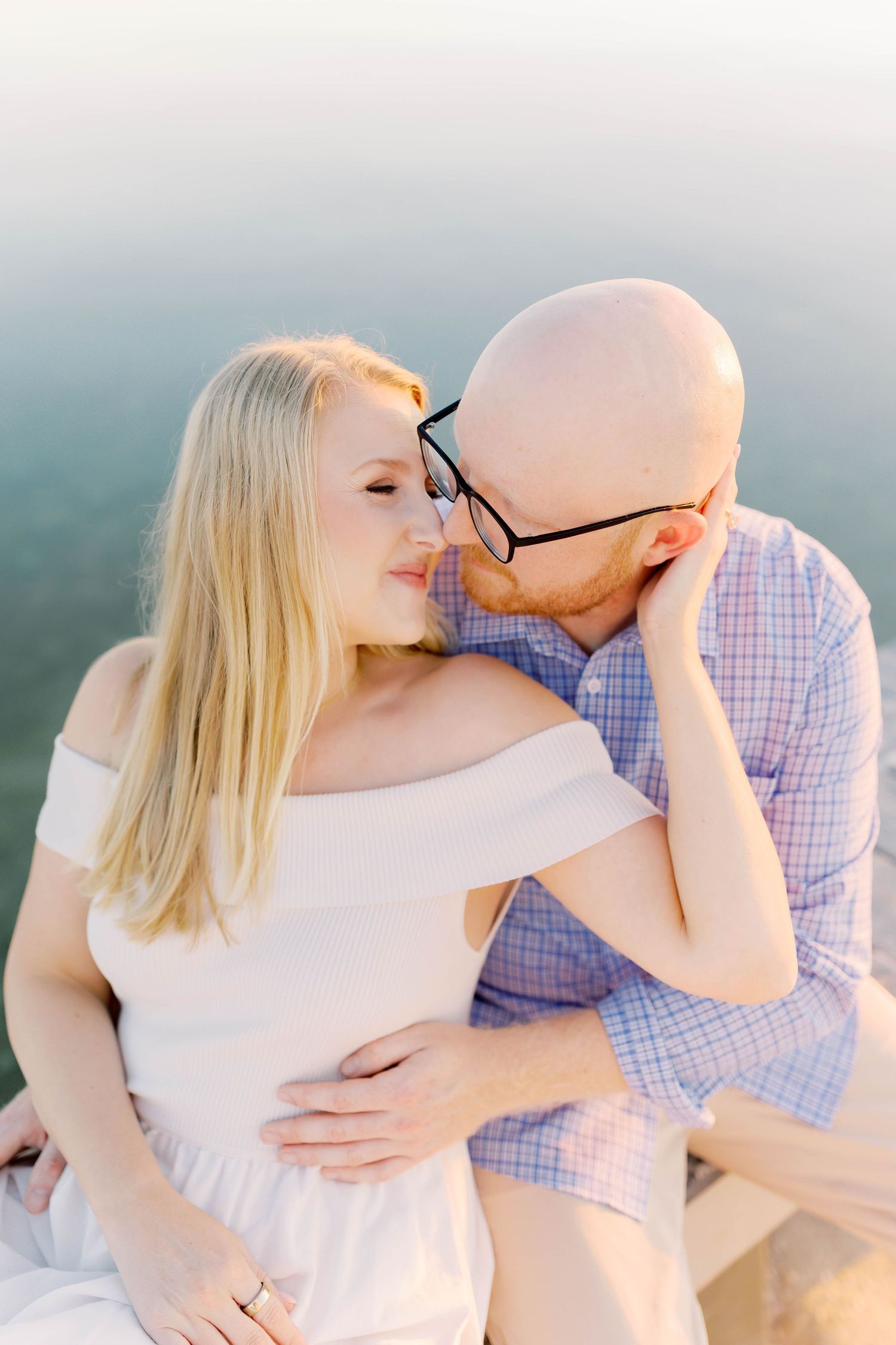 Romantic couple in white dress and blue checkered shirt share intimate moments against soft pastel backdrop.