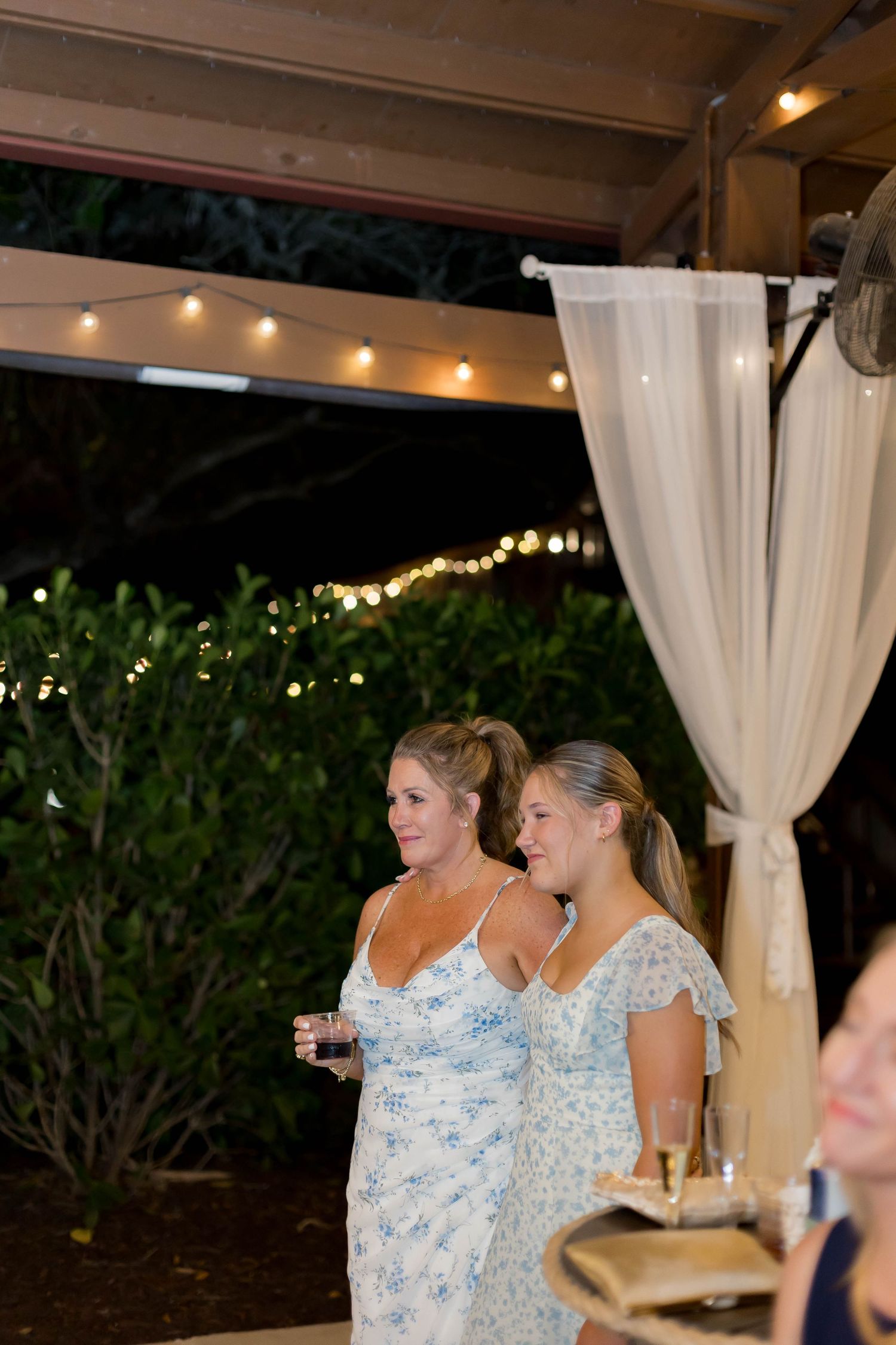 Wedding guests in blue floral dresses enjoying drinks under string lights.