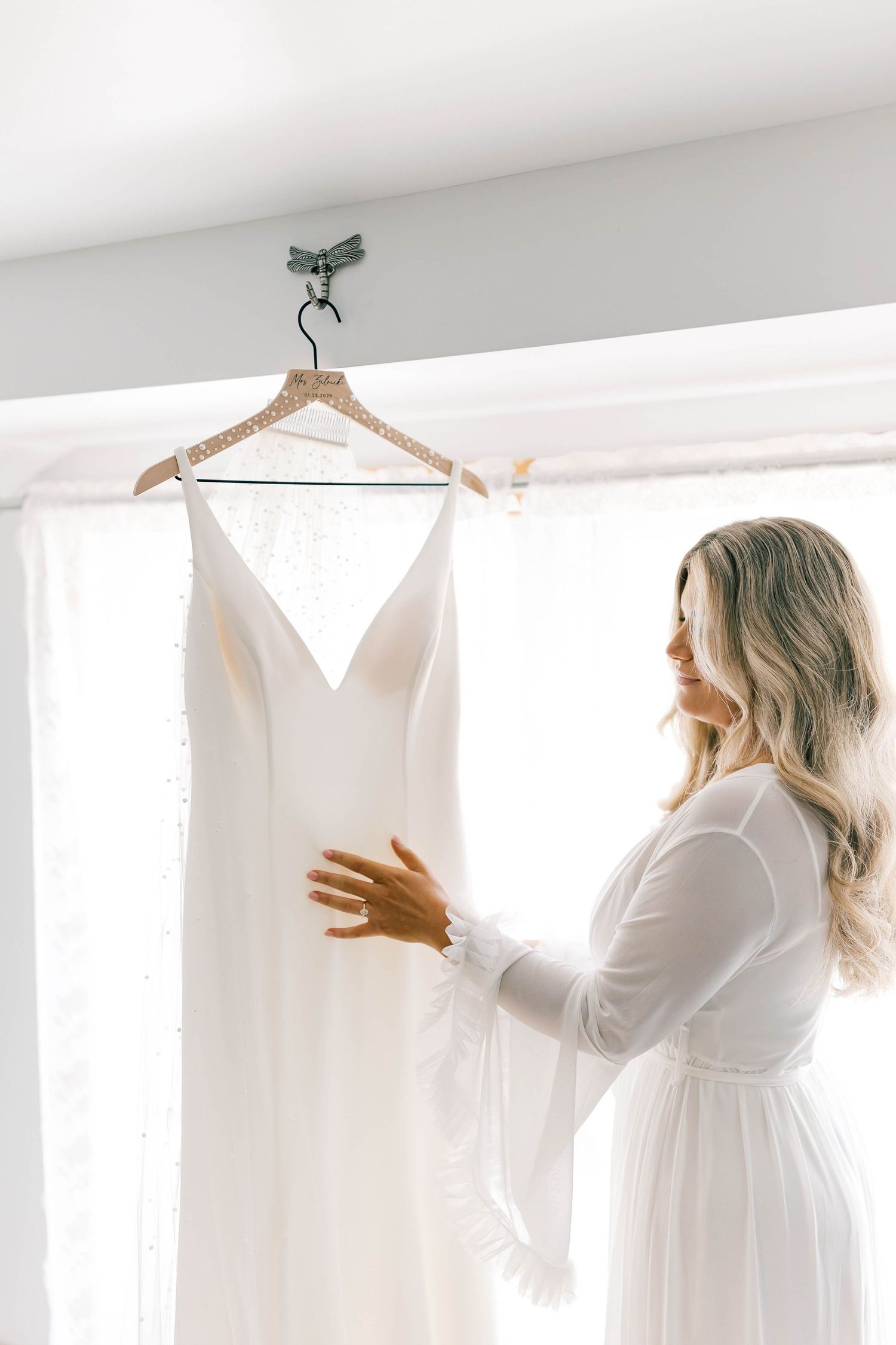 Bright morning wedding preparation scene with white dress hanging in window.